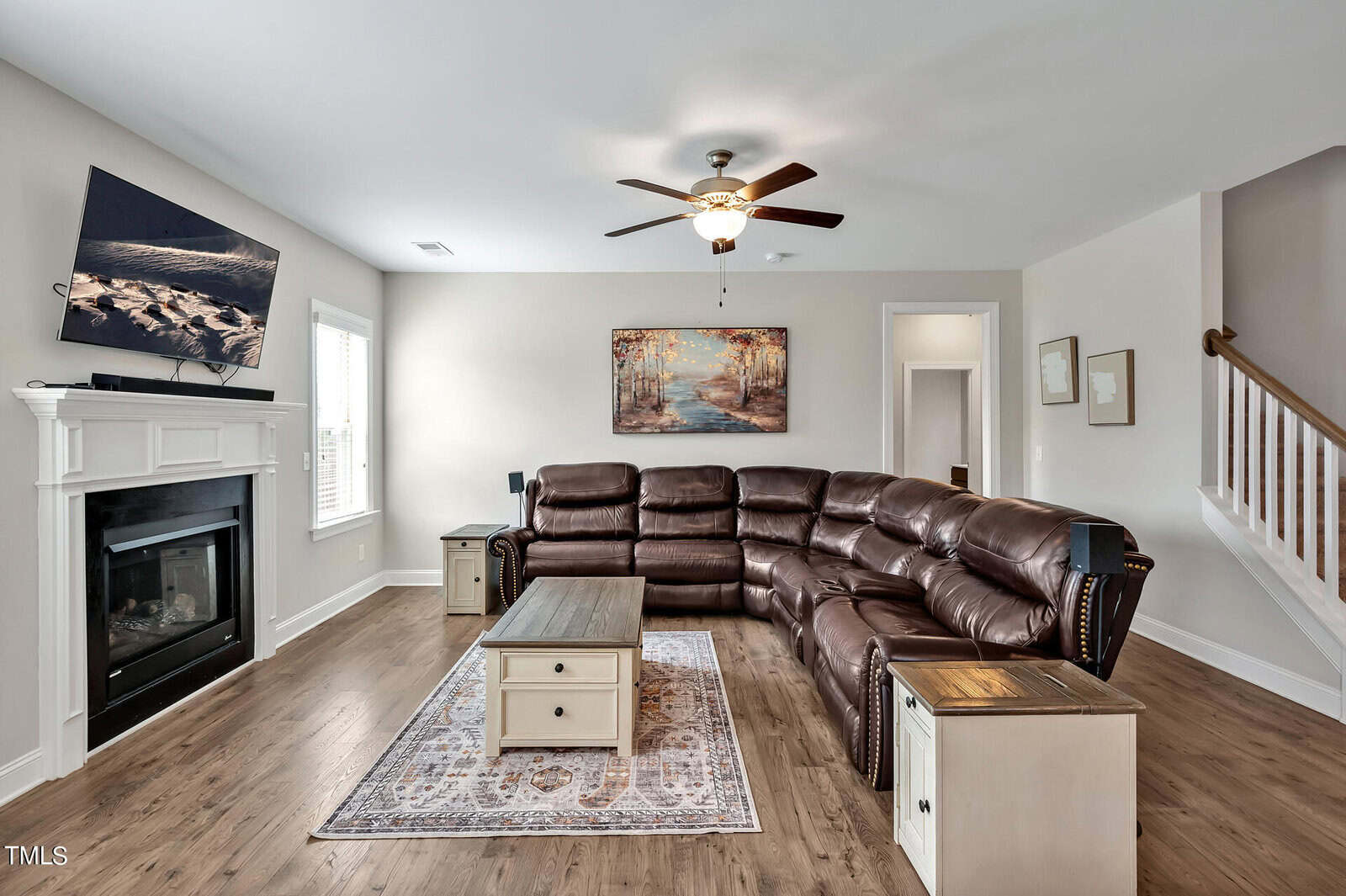 A living room with a brown leather sectional sofa, a ceiling fan, a wall-mounted TV above a fireplace, a coffee table on a rug, and neutral-colored walls decorated with framed artwork.