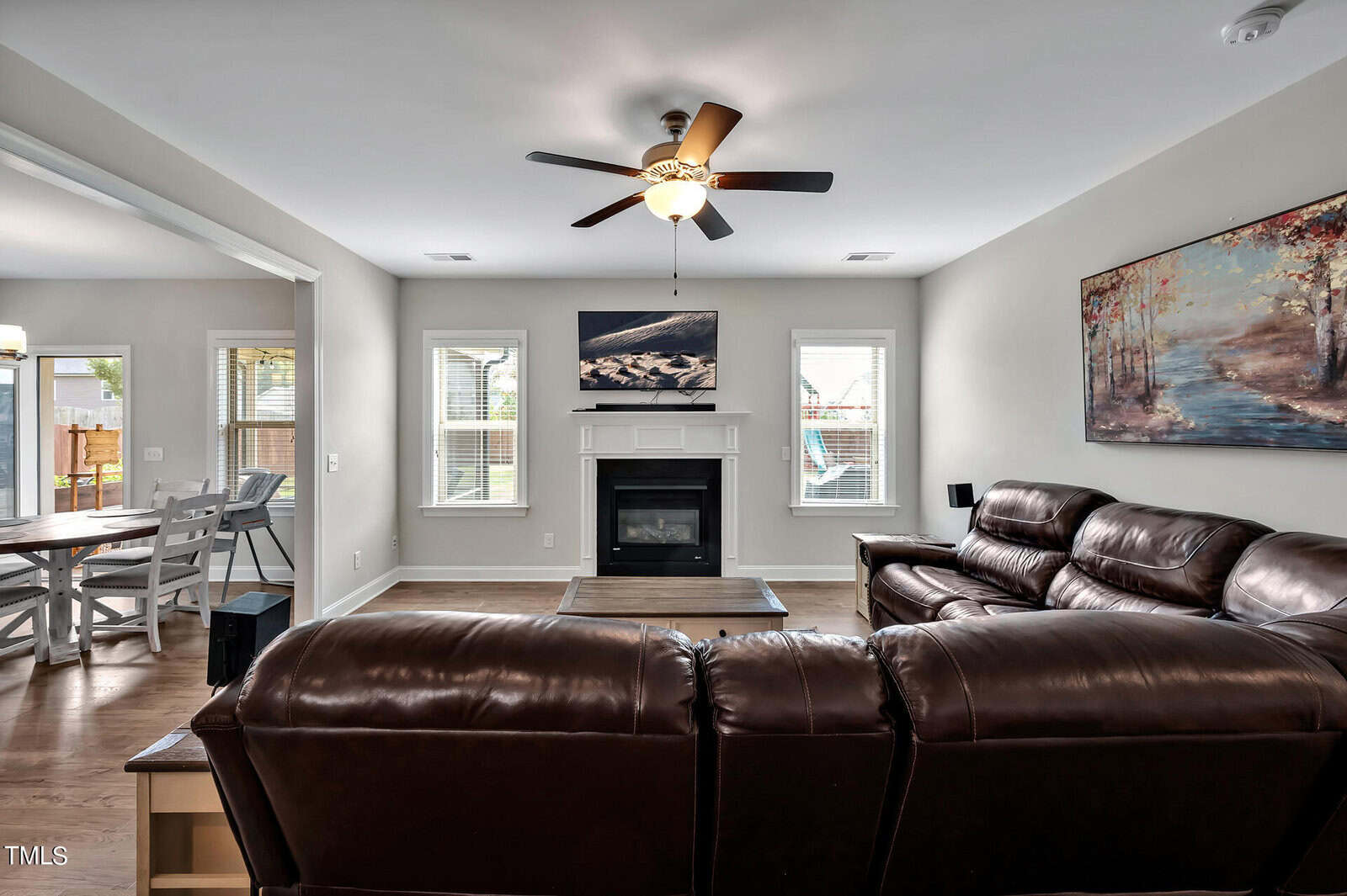 A living room with brown leather sofas, a wooden coffee table, a ceiling fan, and a fireplace centered between two windows. Artwork hangs above the fireplace and on the right wall. The dining area is visible to the left.