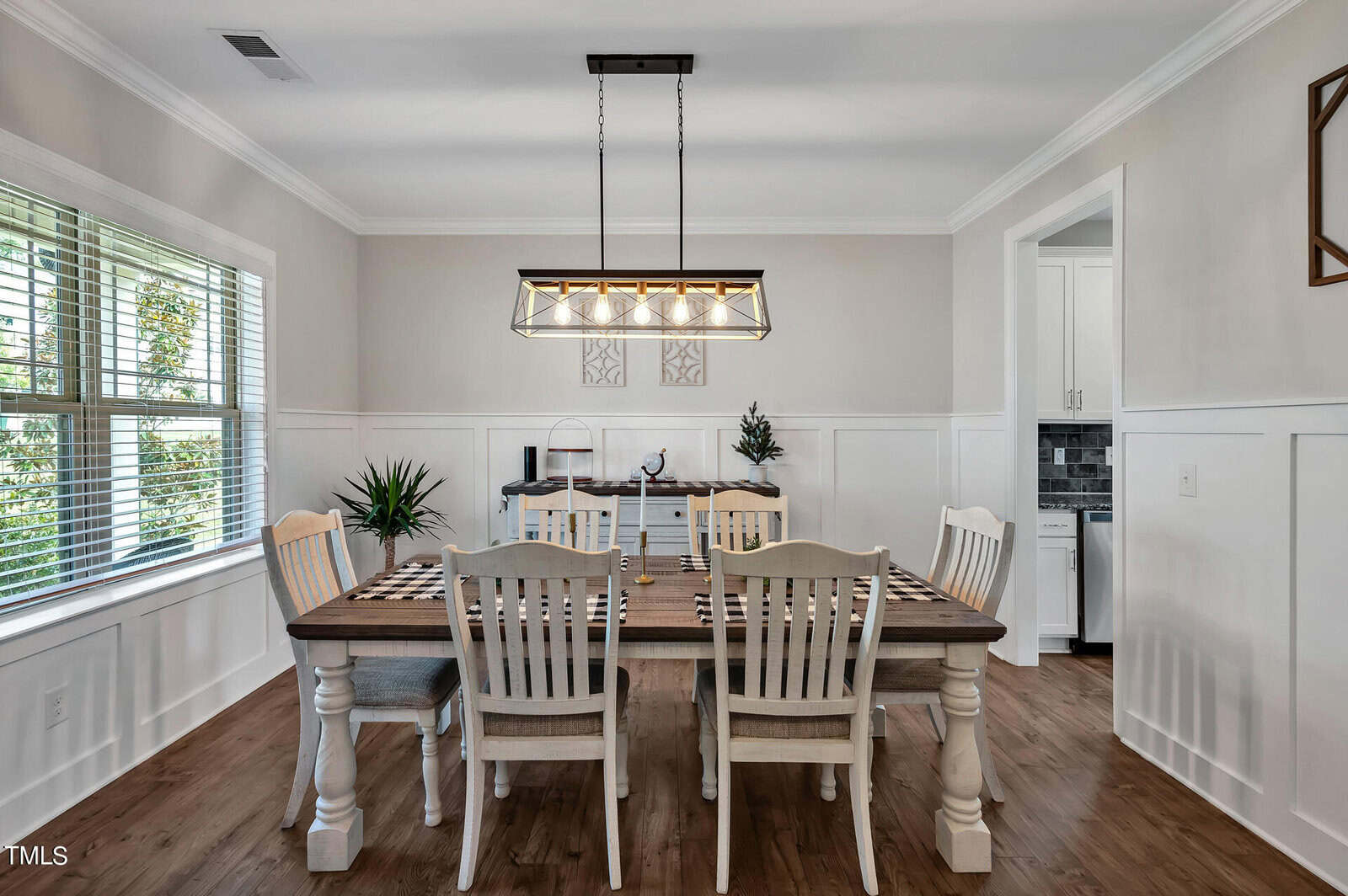A dining room with a wooden table and six chairs, a checkered table runner, a modern rectangular chandelier, hardwood floors, white wainscoting, two potted plants, and a sideboard against the back wall.