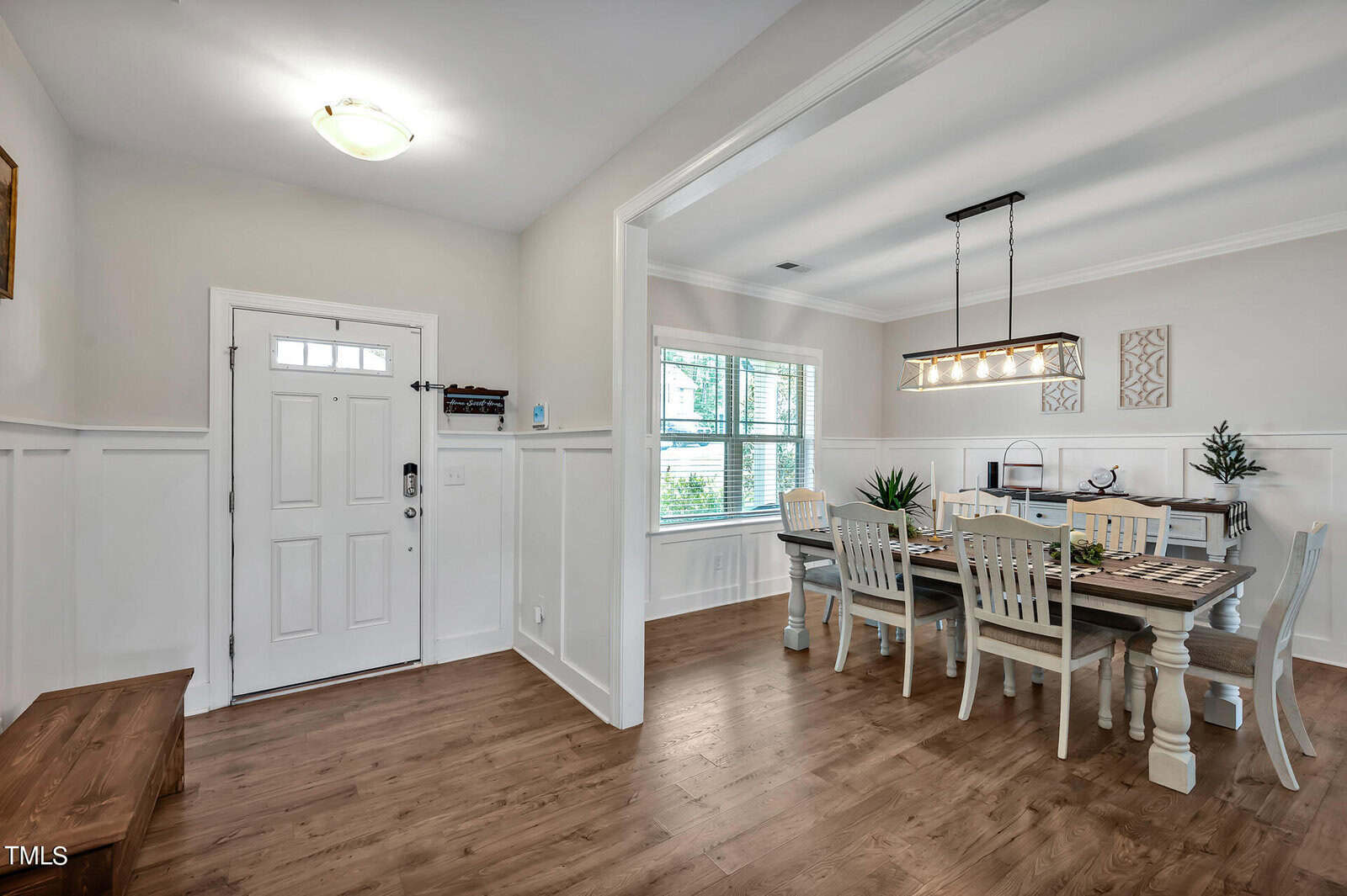 A bright entryway with wood flooring opens into a dining room featuring a rectangular table with six chairs, two large windows, and a modern light fixture above the table. Walls are painted light and decor is minimal.