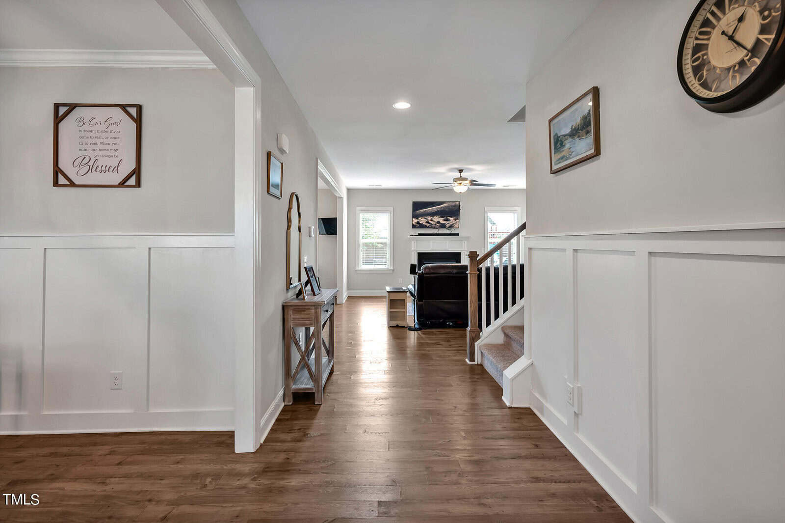 A hallway with wood flooring leads to a living room with a sofa and TV. A staircase with a white railing is on the right, and a console table with a mirror is on the left. Walls are light-colored with wainscoting.
