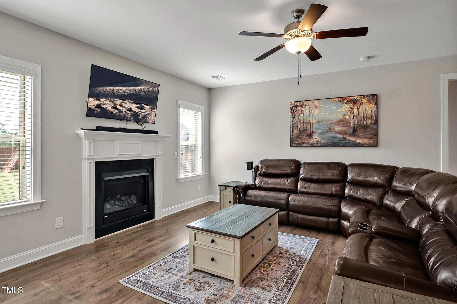 A cozy living room with brown leather sectional, white fireplace, wall-mounted TV, landscape painting, coffee table, hardwood floors, and large windows letting in natural light.