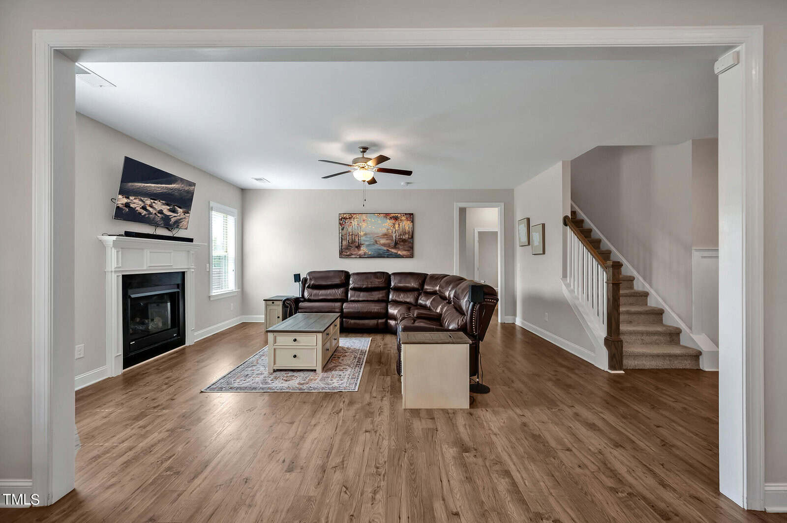 A spacious living room with wood flooring, a sectional sofa, a coffee table, a fireplace with a TV above, wall art, and a staircase leading upstairs. Large windows let in natural light.