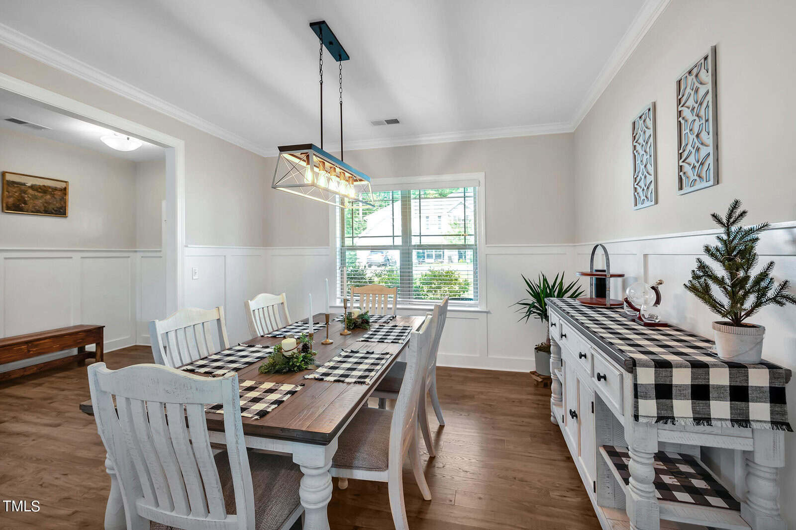 A dining room with a wooden table set for six, white chairs, a black-and-white checkered table runner, matching sideboard, wall art, and a large window letting in natural light.