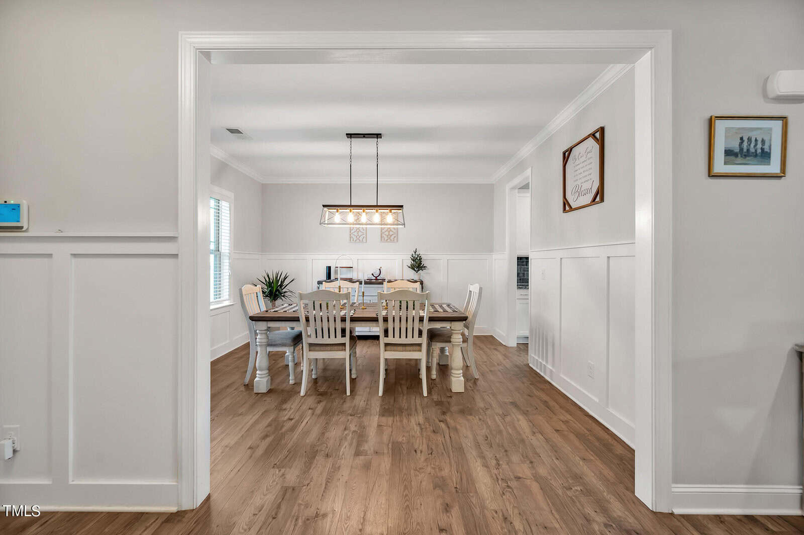 A dining room with a wooden table, six chairs, pendant lighting above the table, wood flooring, white wainscoting on the walls, and framed artwork hanging on the walls.
