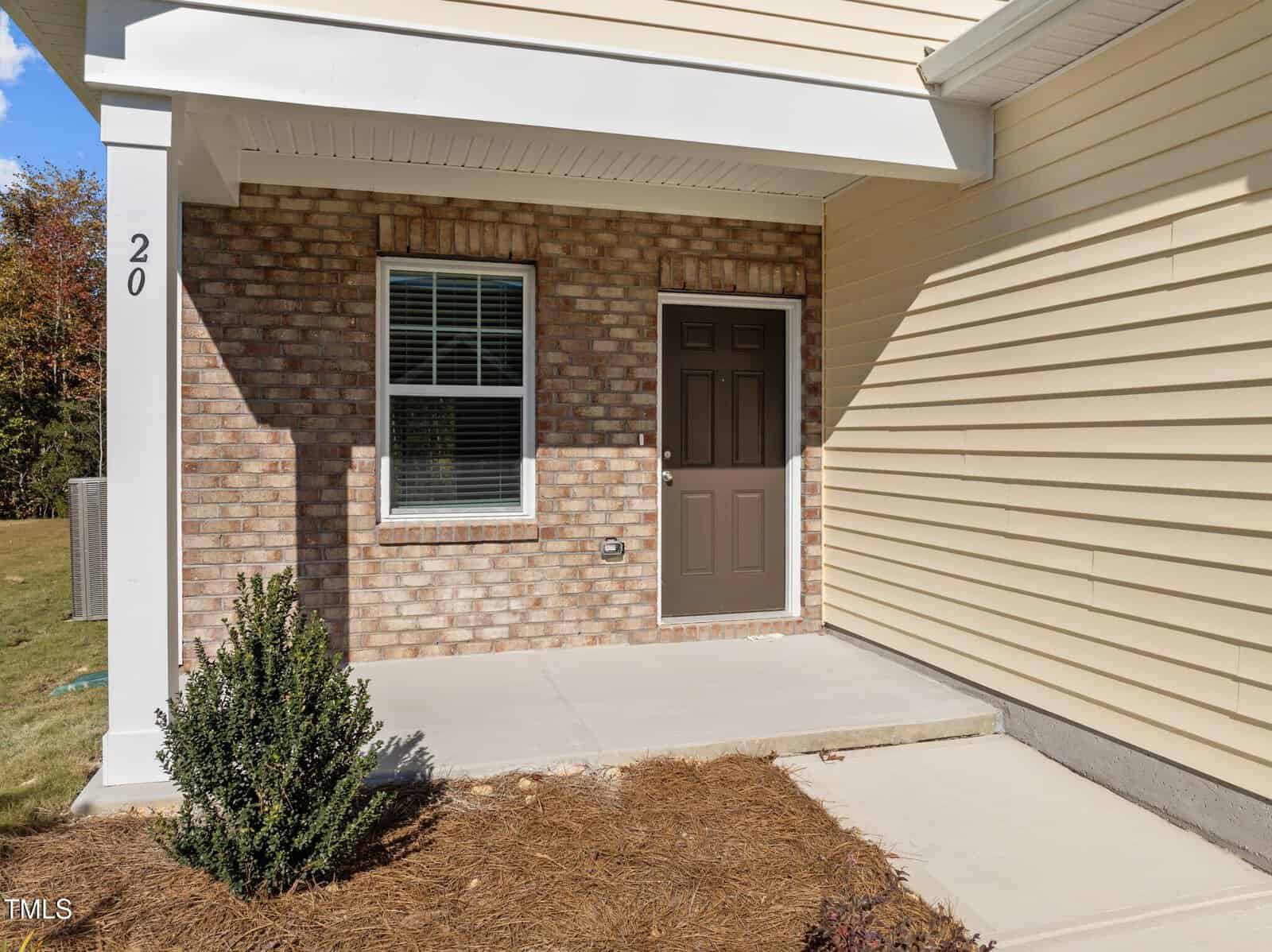 A small covered front porch with a concrete floor, beige siding, brick wall, a window with white trim, a brown door, and a white post labeled "20". There is a shrub and mulch around the porch.