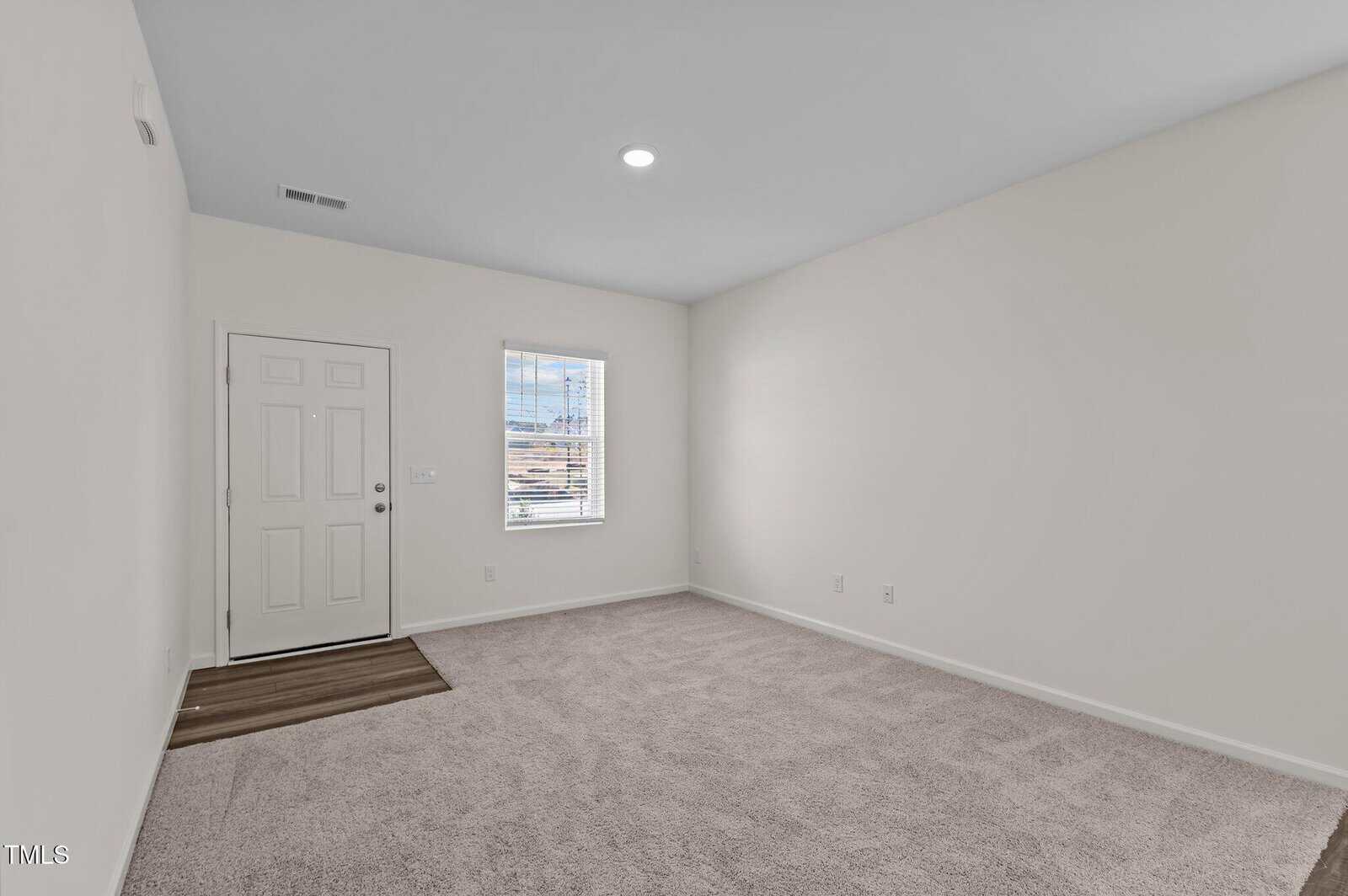 A small empty room with beige carpet, white walls, a white door, a window with blinds, and a section of wood-look flooring near the entrance. A single ceiling light is on.