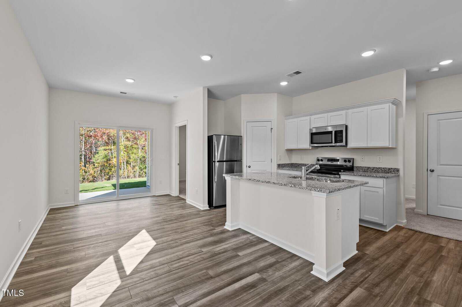 Open-concept kitchen and living area with stainless steel appliances, white cabinets, a granite island, wood-style flooring, recessed lighting, and glass doors leading to a patio with a view of trees.