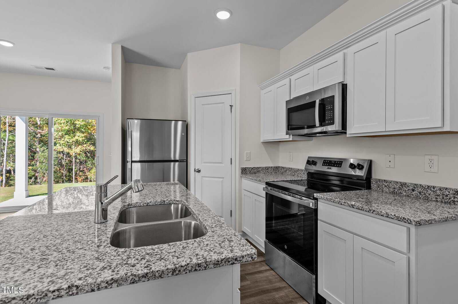 A modern kitchen with white cabinets, granite countertops, a stainless steel refrigerator, microwave, and oven, a double sink, and sliding glass doors leading outside. The floor is wood-look and walls are light-colored.
