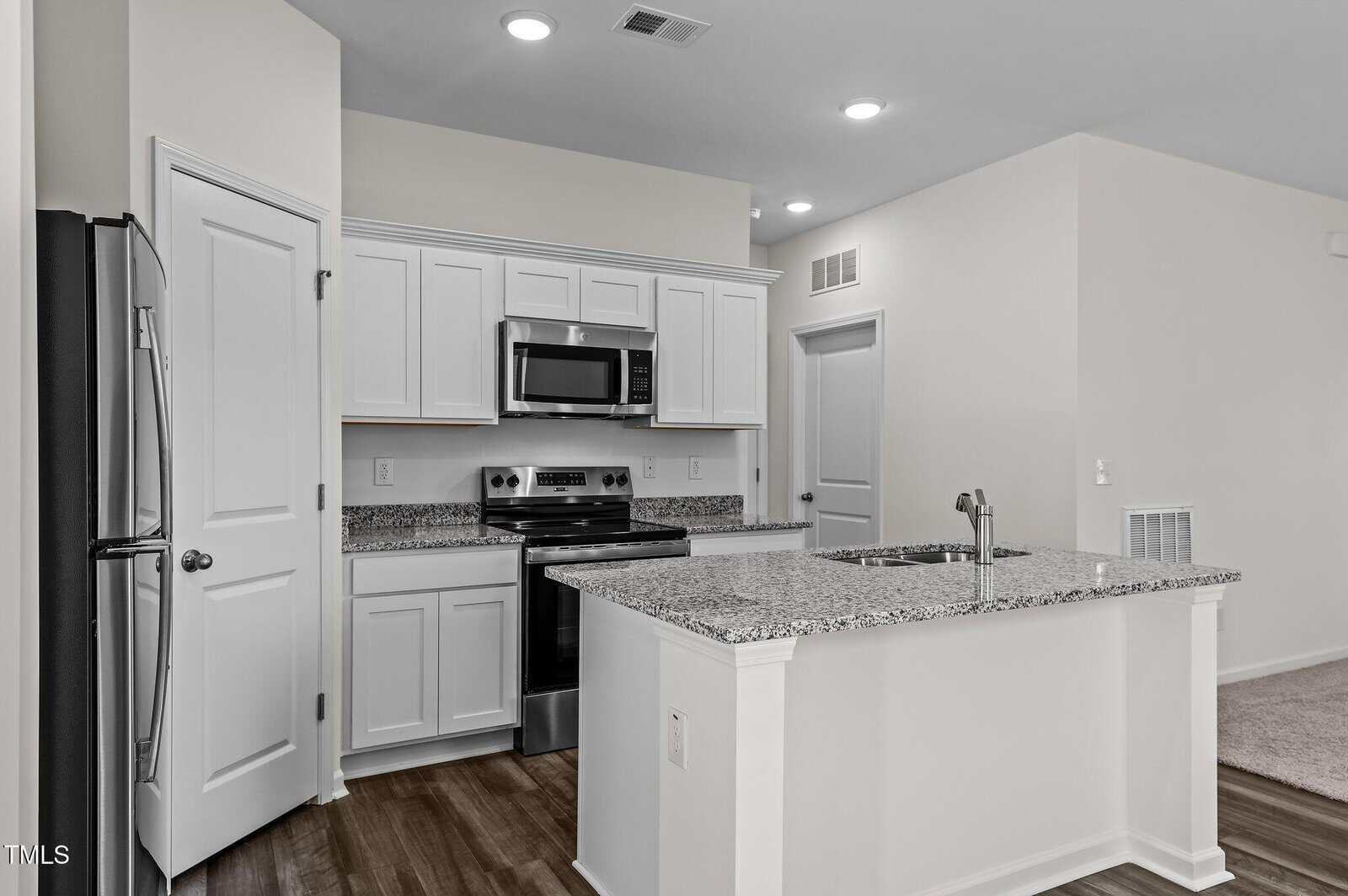 Modern kitchen with white cabinets, granite countertops, stainless steel appliances, an island with a sink, and wood flooring. The walls are light-colored and the room is well-lit with recessed ceiling lights.