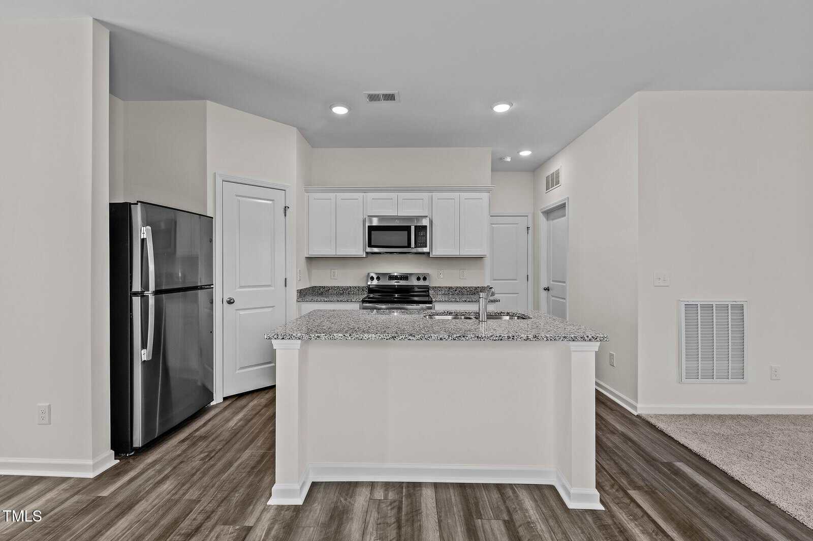 A modern kitchen with white cabinets, stainless steel appliances, granite countertops, and wood-style flooring. There is an island with a sink in the center and a refrigerator to the left. The walls are painted a light color.