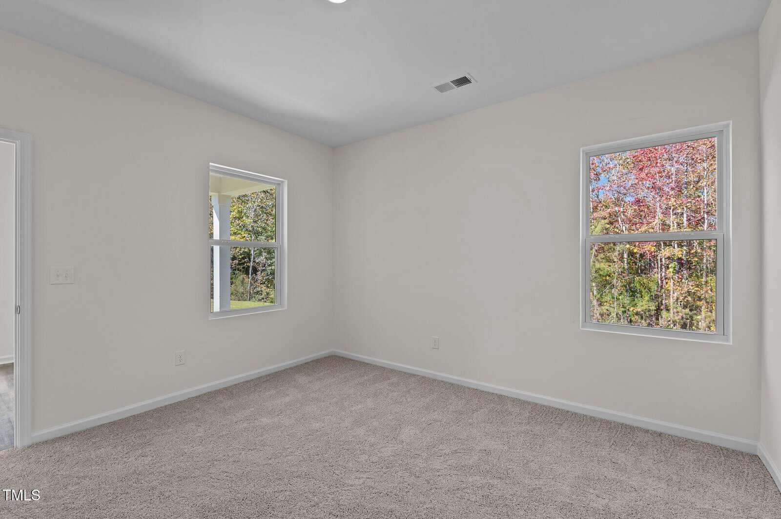Empty room with beige carpet, white walls, two windows showing trees outside, and a doorway to the left.