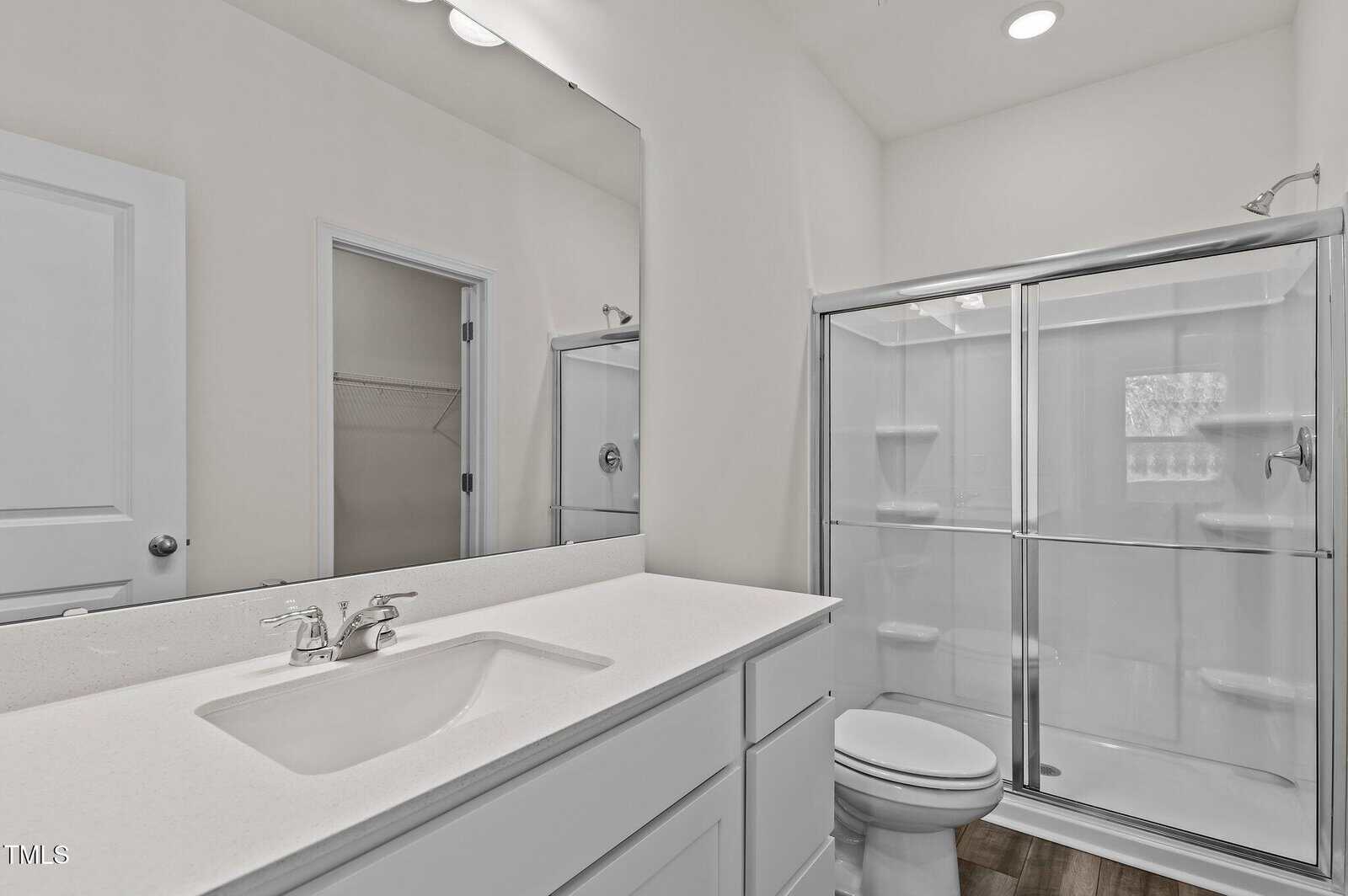Modern bathroom with white cabinets, a large mirror, single sink, toilet, and a glass-enclosed shower. The room has bright lighting and a door leading to an adjacent walk-in closet.