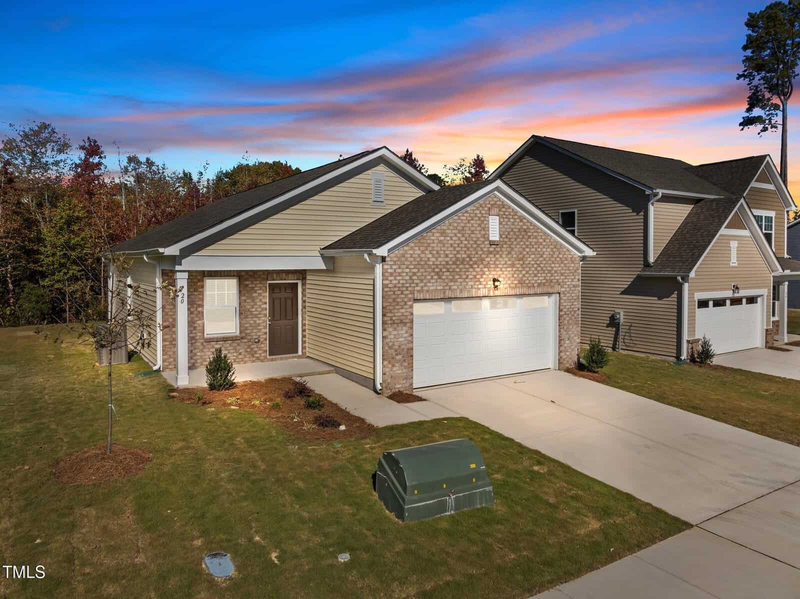 Single-story house with a brick and siding exterior, two-car garage, concrete driveway, small bushes, and a green utility box on the front lawn. The sky shows a colorful sunset. Neighboring house is visible.
