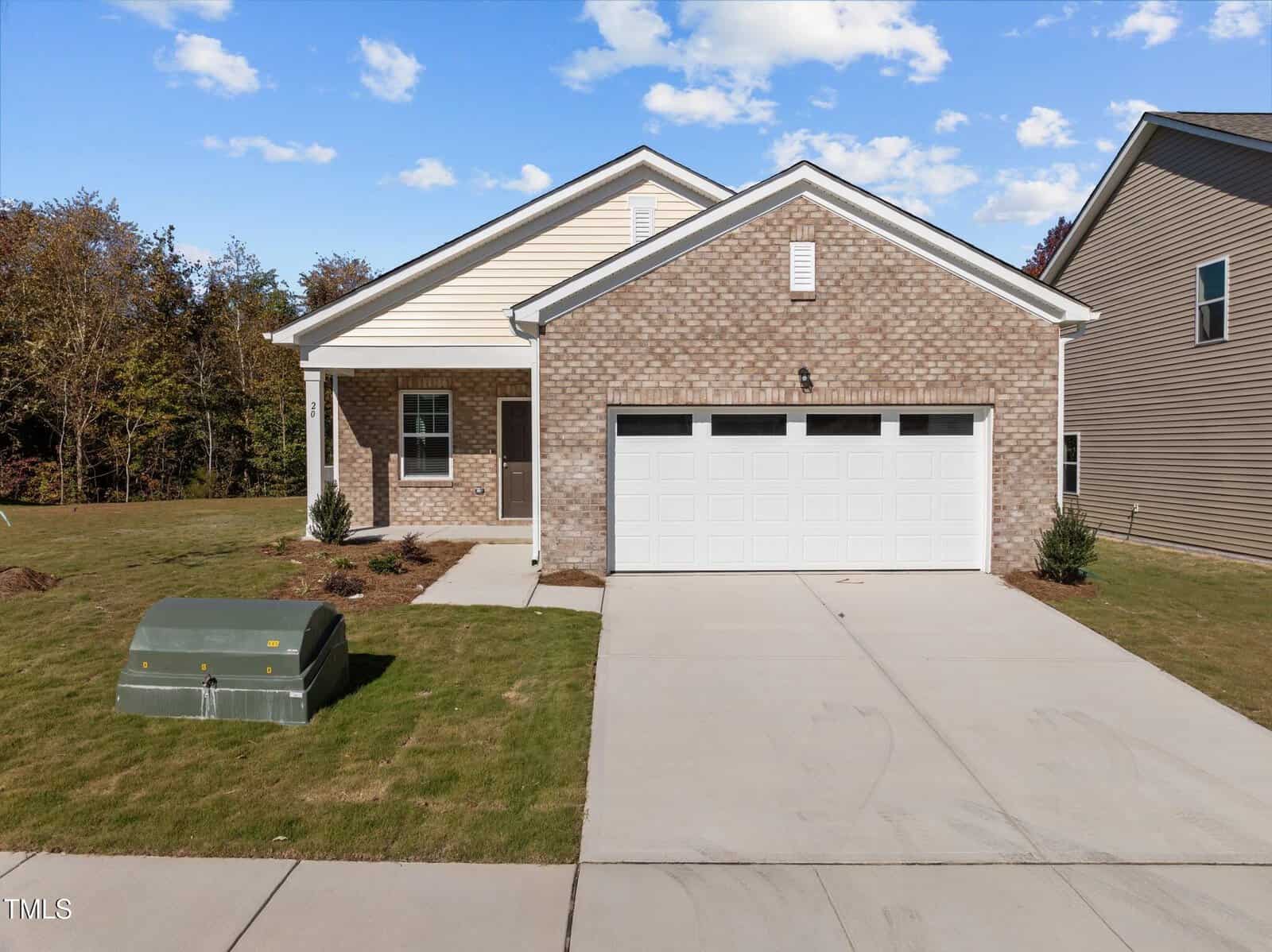 A single-story house with a brick and siding exterior, a two-car garage, and a concrete driveway. There is a small grassy lawn, minimal landscaping, and a green utility box near the sidewalk. Trees are in the background.