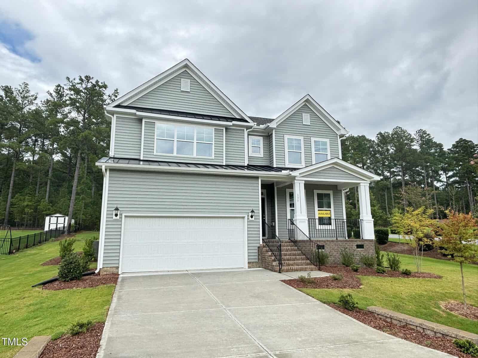 Two-story gray house with white trim and a two-car garage, situated on a grassy lawn with mulch, shrubs, and a paved driveway at 135 Green Haven Boulevard in Youngsville. Forested area and cloudy sky in the background.