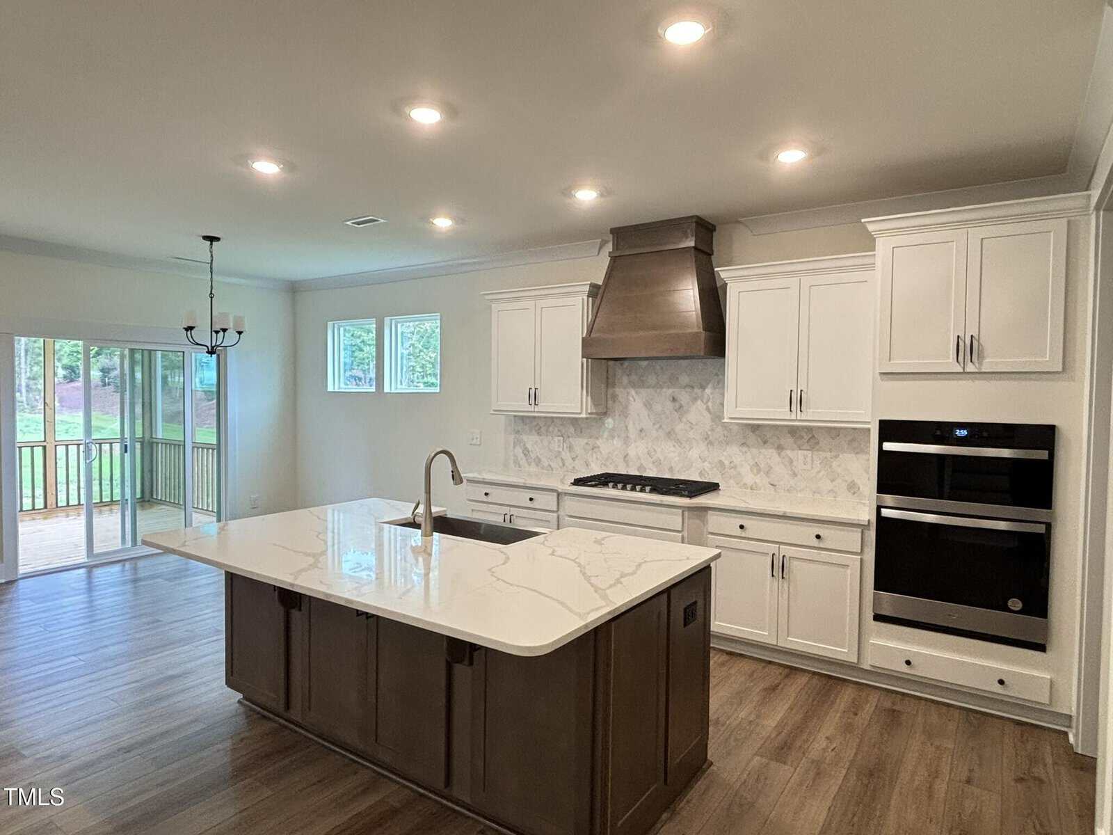 Modern kitchen with white cabinets, stainless steel appliances, marble backsplash, and a large island with sink. Hardwood flooring and recessed lighting. Sliding doors and windows provide natural light and a view of a porch and backyard.