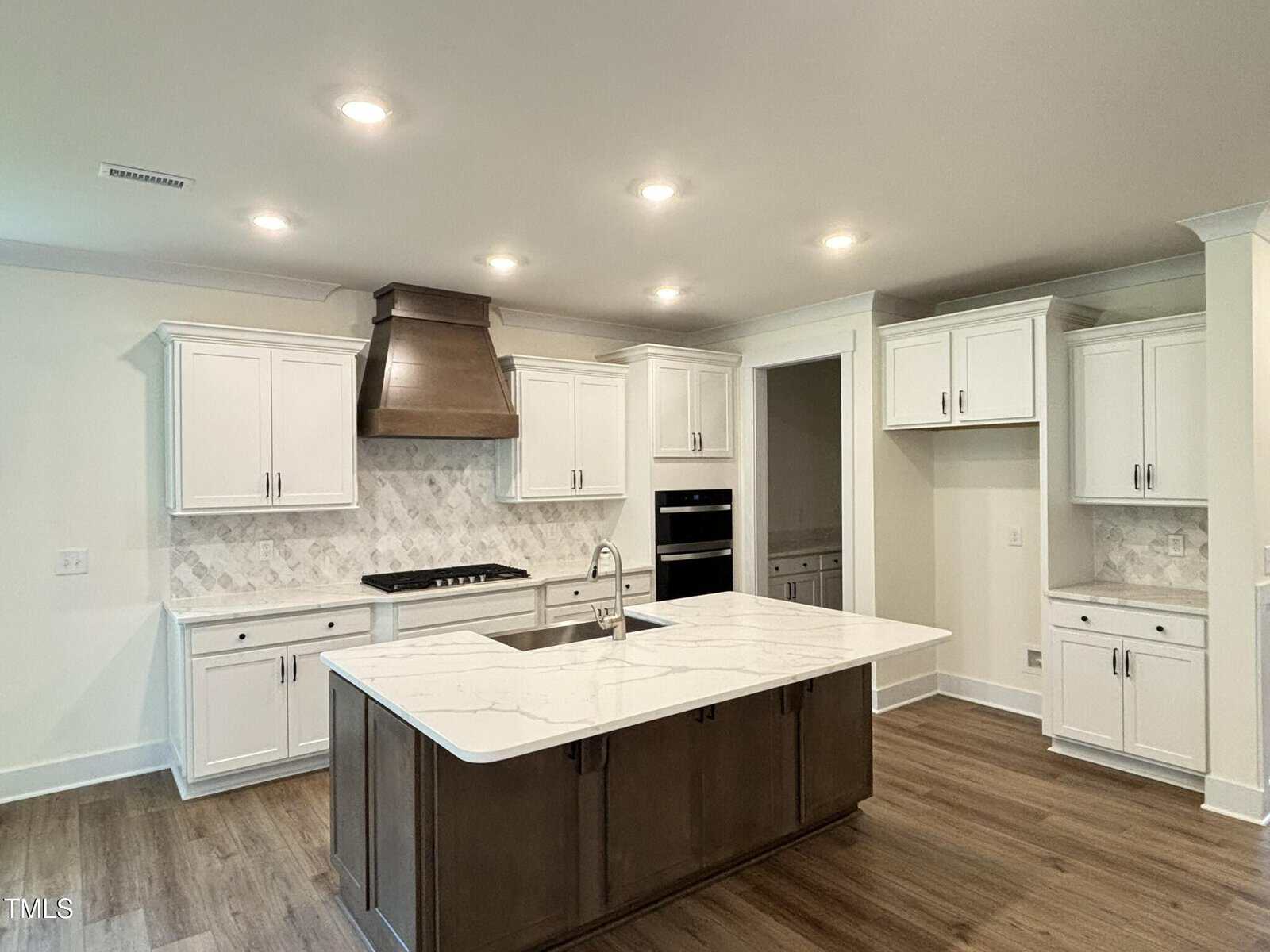 Modern kitchen with a large center island, white cabinetry, stainless steel appliances, wood floors, marble countertops, and a tile backsplash. Recessed ceiling lights illuminate the room. No people are present.