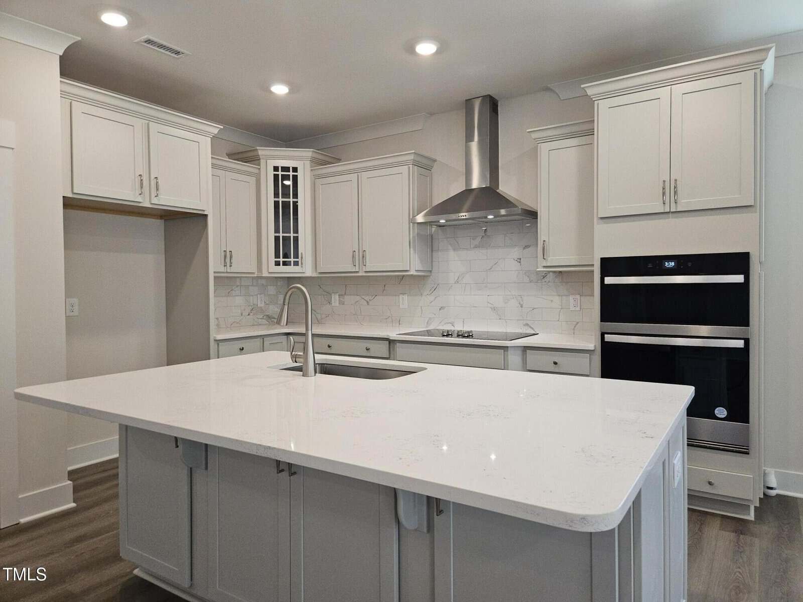 Modern kitchen with white cabinets, stainless steel appliances, a vent hood, double wall ovens, marble backsplash, and a large white island with a built-in sink and gooseneck faucet. Light wood flooring throughout.