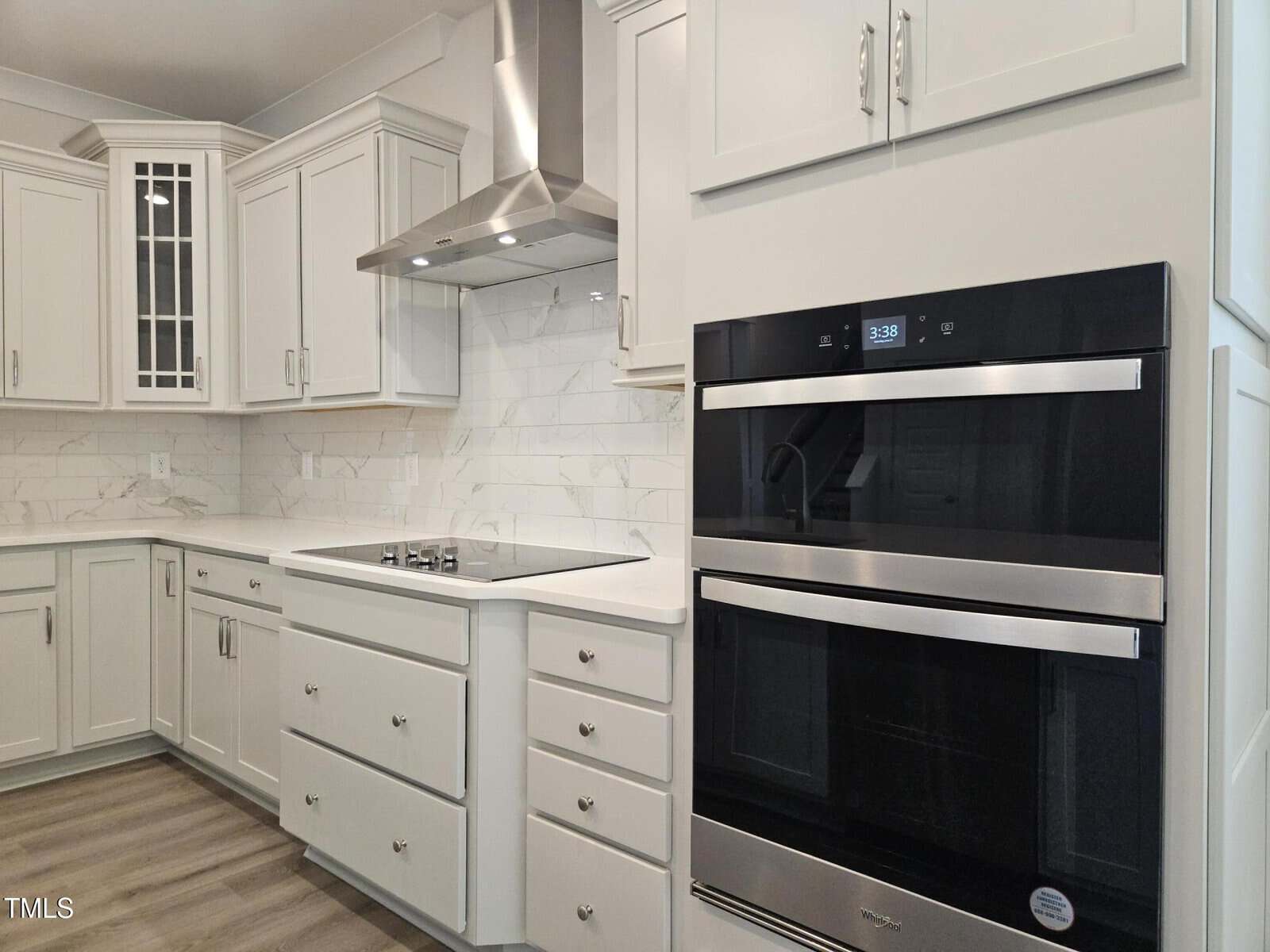 Modern kitchen with white cabinets, stainless steel oven, glass cooktop, and a range hood. The backsplash is white tile with a marble pattern, and the floor is wood. Upper cabinets have glass doors.