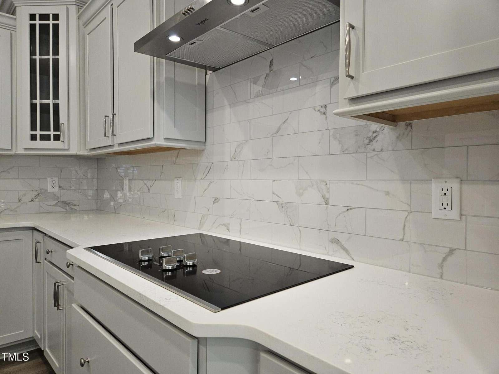 Modern kitchen with a white marble tile backsplash, white countertops, a black glass cooktop with knobs, and light gray cabinets with stainless steel handles.