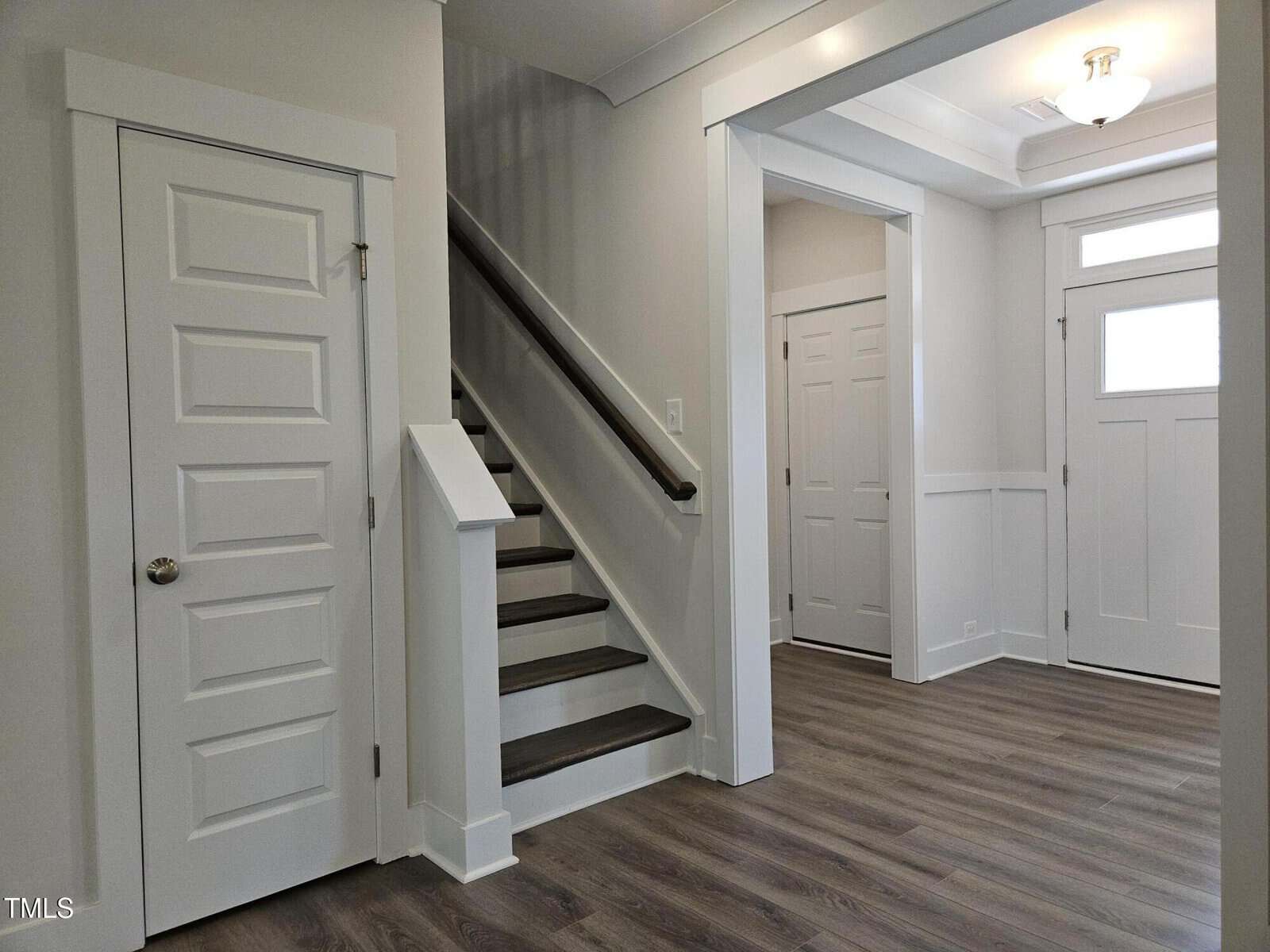 Modern entryway with wood-look vinyl flooring, white paneled doors, a staircase with dark handrail, and natural light from a window above the front door. Clean, neutral color palette.