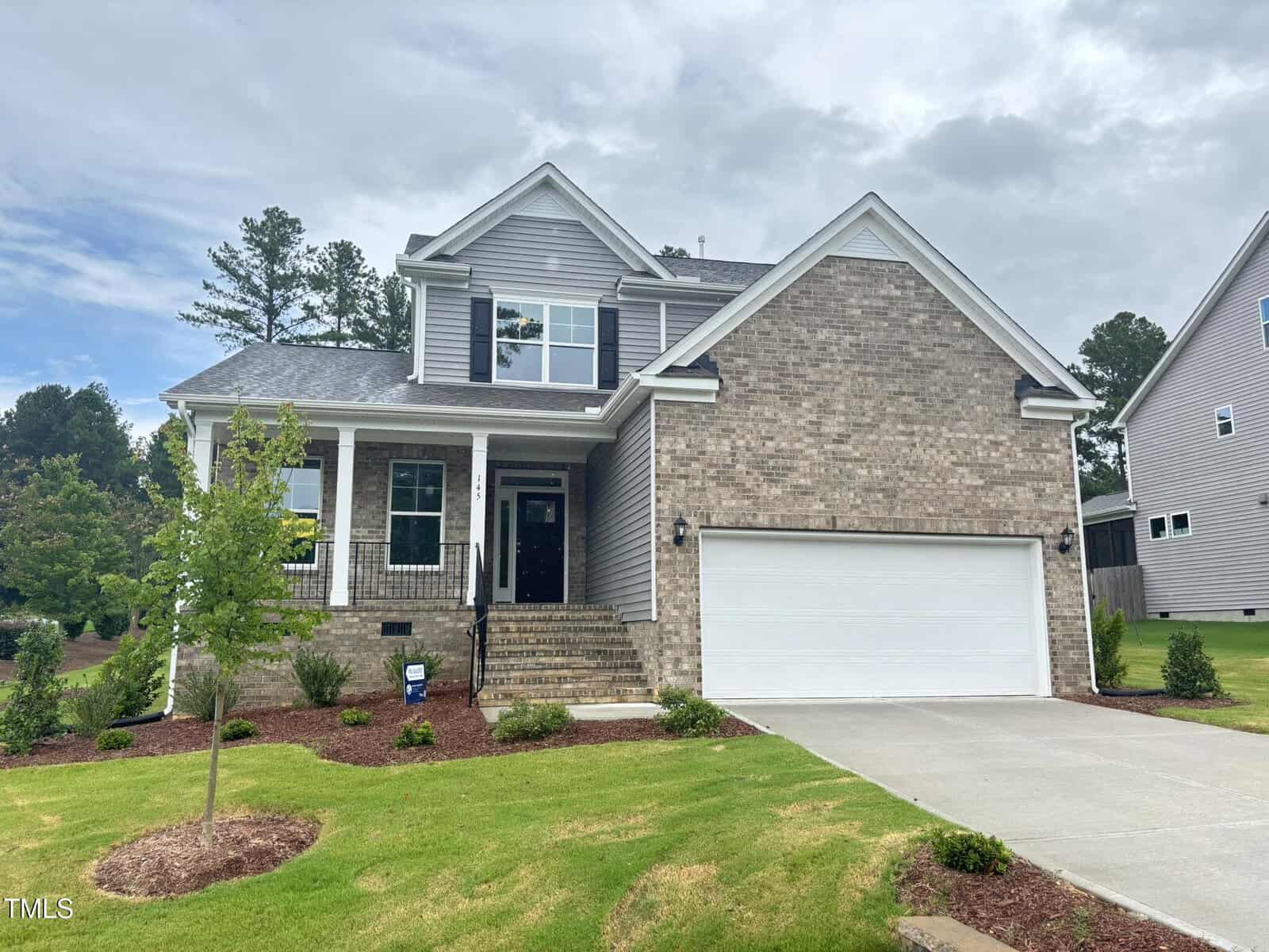 Two-story suburban house with brick and gray siding, front porch, double garage, and neatly landscaped yard with a small tree, grass, and bushes on a cloudy day, located at 145 Green Haven Boulevard in Youngsville.