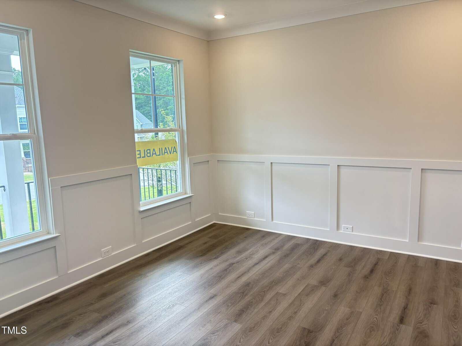 Bright, empty room with light beige walls, white trim, large windows, and wood-style flooring. Recessed ceiling lights are on, and a yellow “Available” sign is visible through the window.