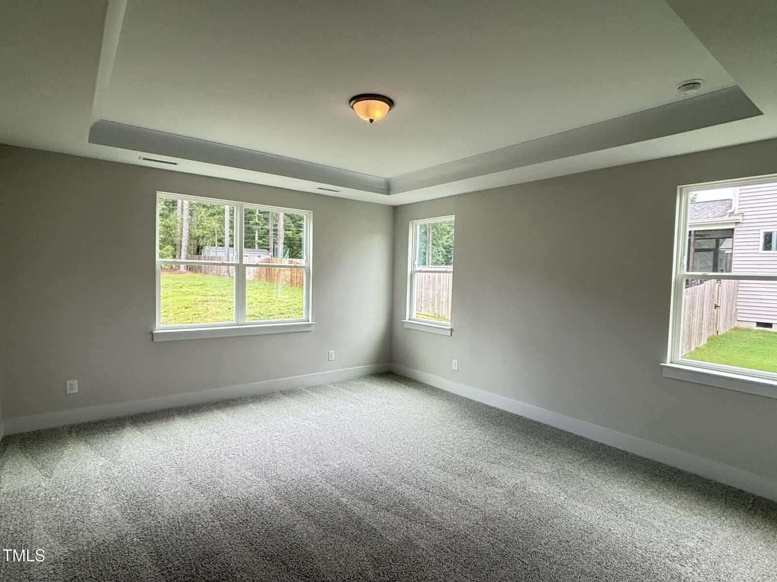 A bright, empty room with light gray walls, a tray ceiling with a central light fixture, and three large windows offering views of a grassy backyard and neighboring house. The room has carpeted flooring.