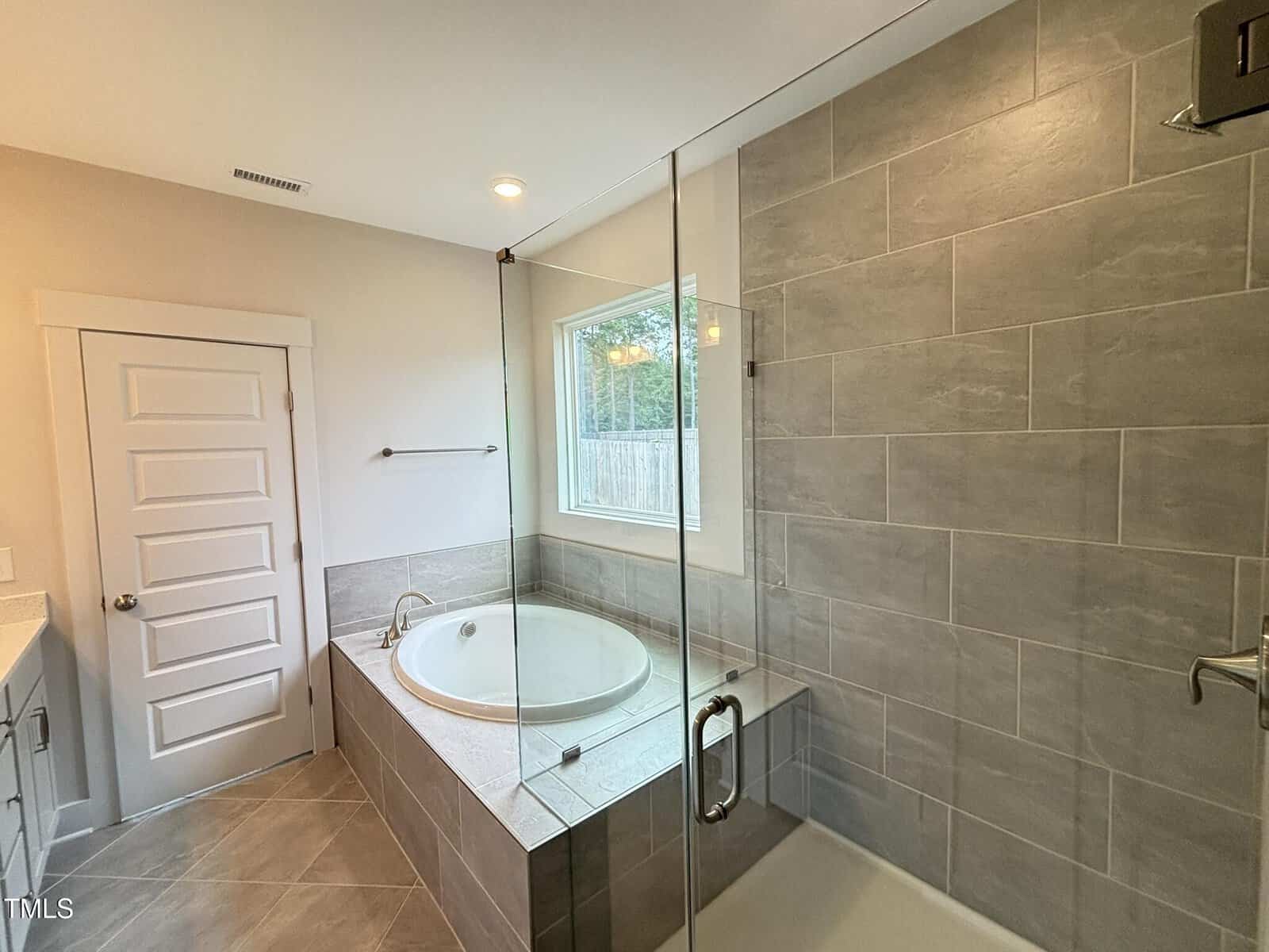 Modern bathroom with a large tiled shower, a built-in bathtub next to a window, a towel bar on the wall, and a white door leading out of the room. Neutral tones and clean lines create a bright, spacious feel.
