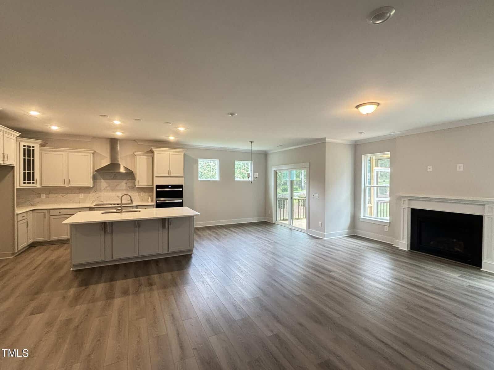 Spacious open-concept kitchen and living area with light wood flooring, white cabinets, large island, double oven, fireplace, and multiple windows letting in natural light. Glass doors lead to a wooden deck outside.