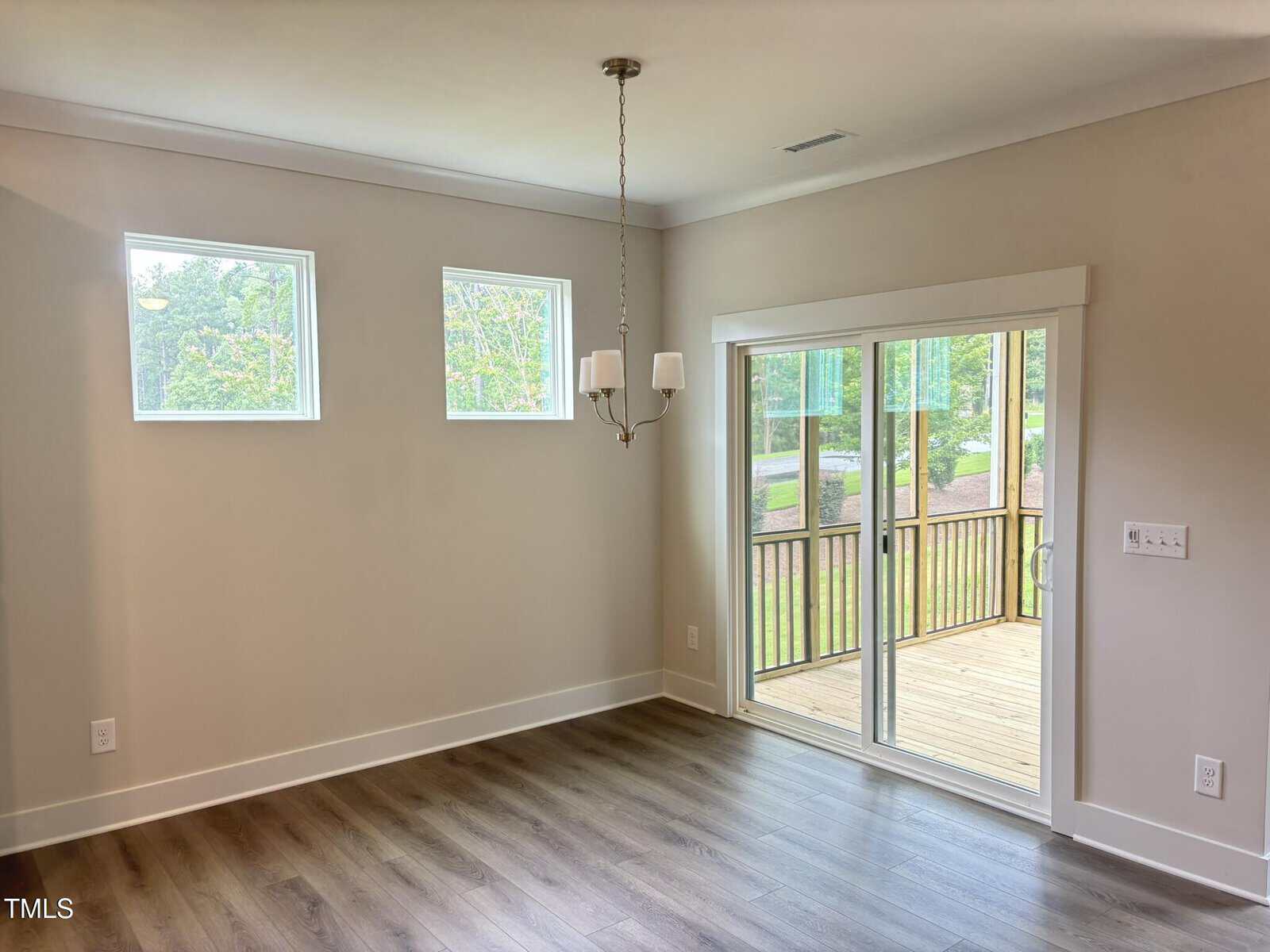 A bright, empty room with light-colored walls, two high rectangular windows, wood flooring, a ceiling light fixture, and sliding glass doors leading to a screened porch and a view of greenery outside.