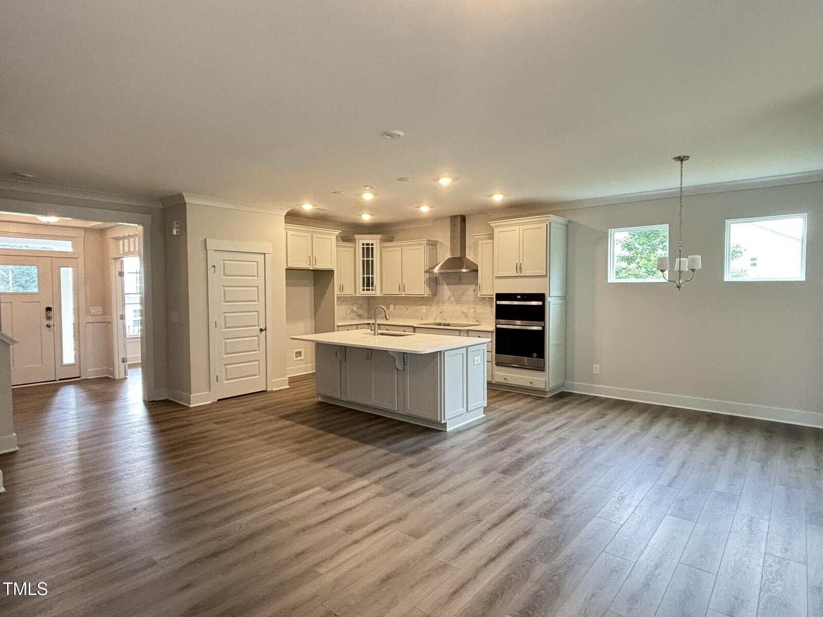 Spacious modern kitchen with light gray cabinets, center island, built-in oven, stainless steel appliances, and wood-look flooring. Open layout with large windows and dining area, leading to entryway.