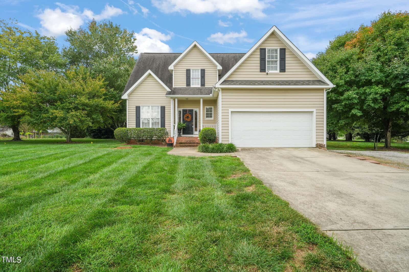 Nestled under a blue sky in Youngsville, the beige two-story house at 192 Jonah Davis Road features white trim and a two-car garage. A concrete driveway winds through the well-kept lawn and trees, leading to its inviting entrance.