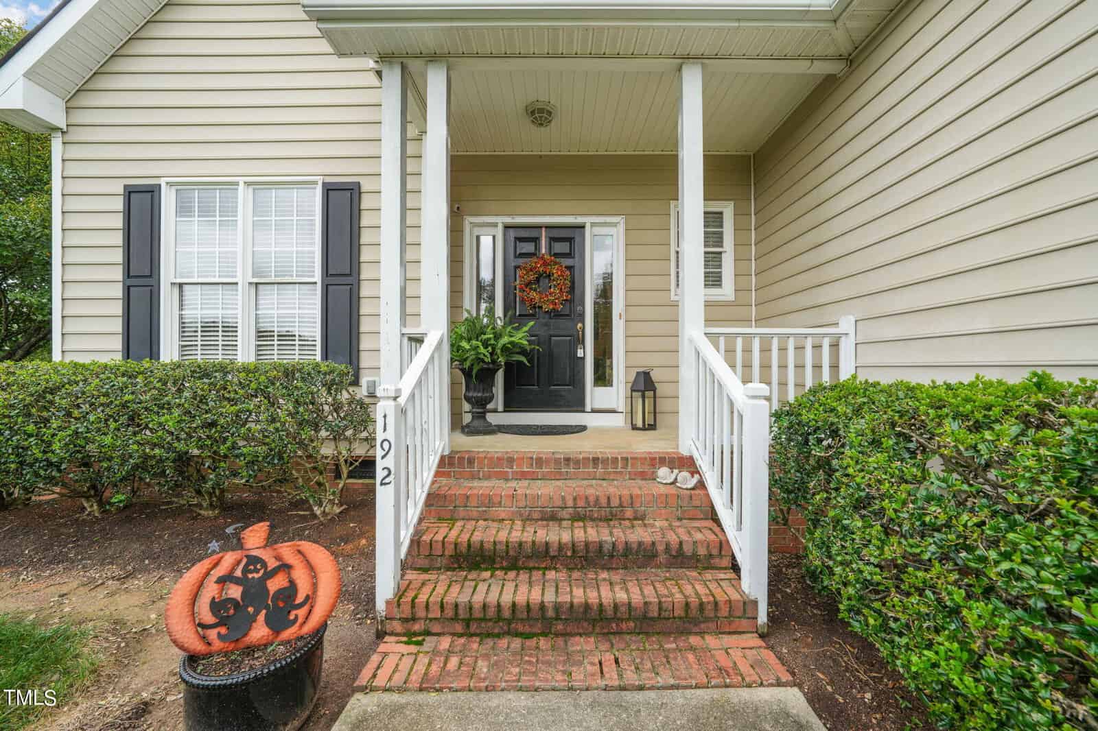 Front porch of a house with light beige siding and black shutters, featuring a black door adorned with an autumn wreath. The steps are red brick, and there's a decorative pumpkin with a cat design near the walkway.