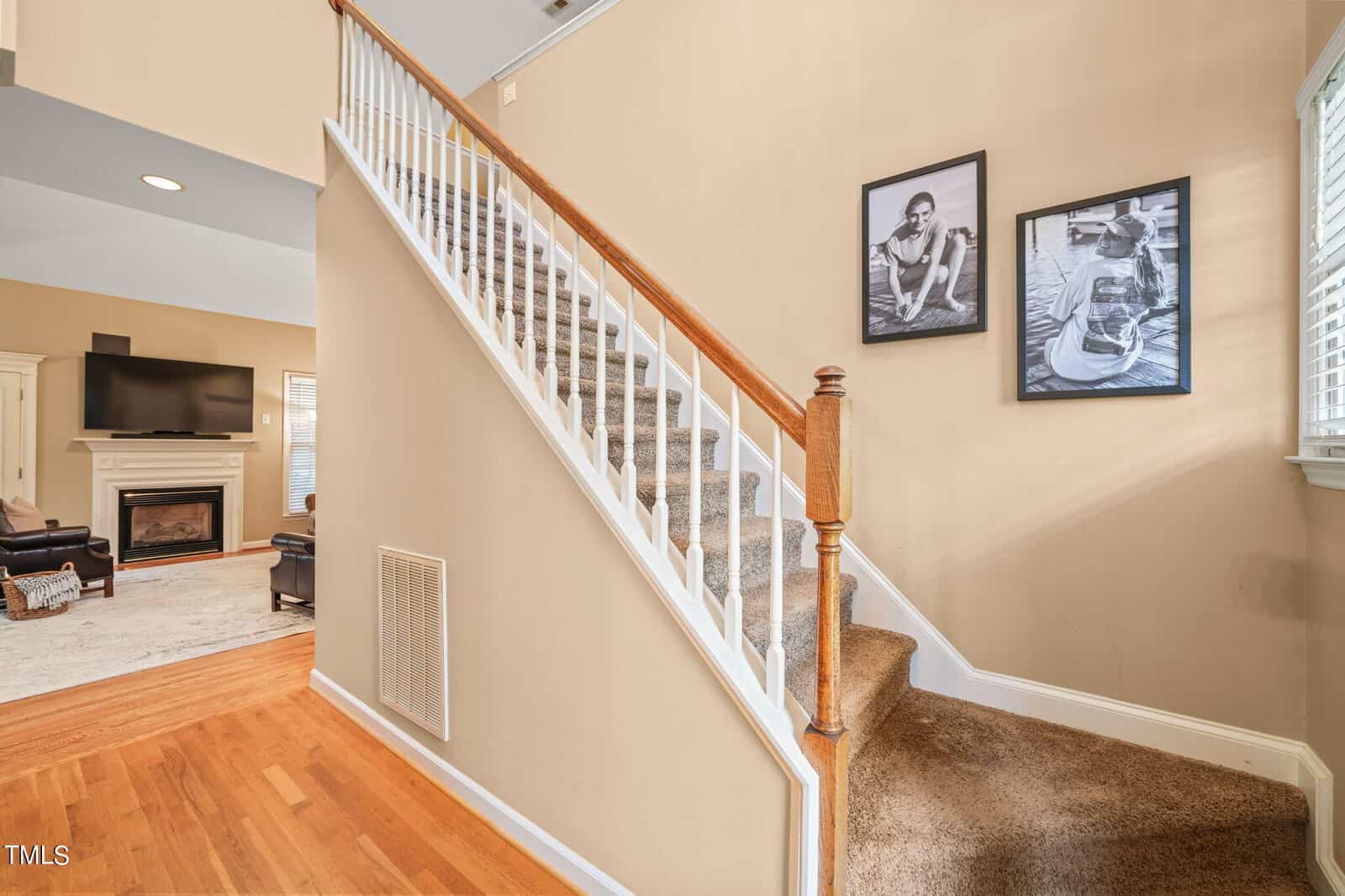 A cozy living room with a carpeted staircase leading upwards. Two framed photos hang on the beige wall beside the stairs. The room has hardwood floors, a flat-screen TV mounted above a fireplace, and a black leather chair in the corner.