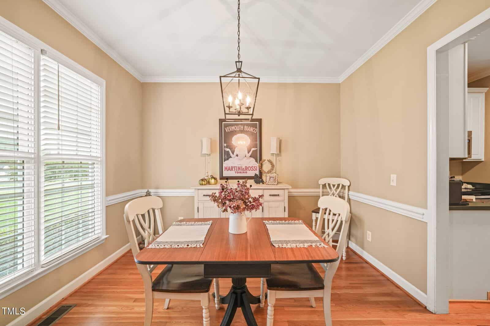 A dining room with a wooden table and four chairs. A vase with flowers is on the table. Beige walls feature a framed poster. A lantern-style light fixture hangs above. Large windows provide natural light, and there's an adjacent open doorway to the kitchen.