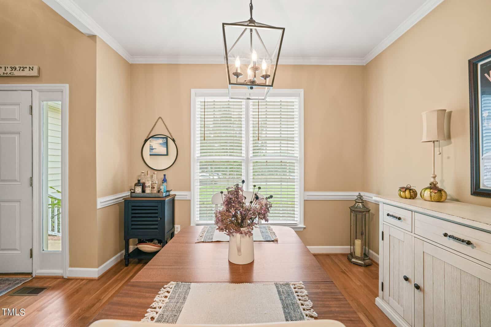 A dining room with a wooden table set with a floral centerpiece. A chandelier hangs overhead. The room features beige walls, a large window with blinds, a cabinet with decor items, and a small bar area with a round mirror on the wall.