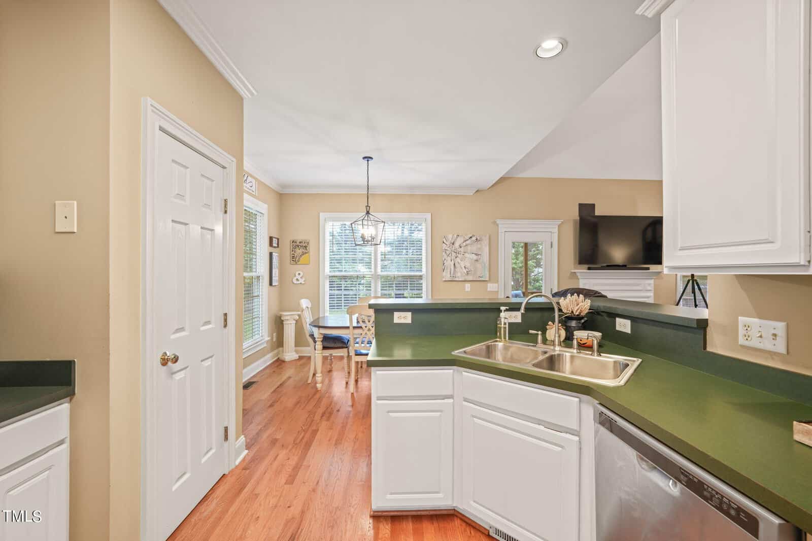 A bright, modern kitchen with white cabinets, green countertops, and stainless steel appliances. There's a double sink, wooden flooring, and a dining area with a round table and chairs next to large windows.