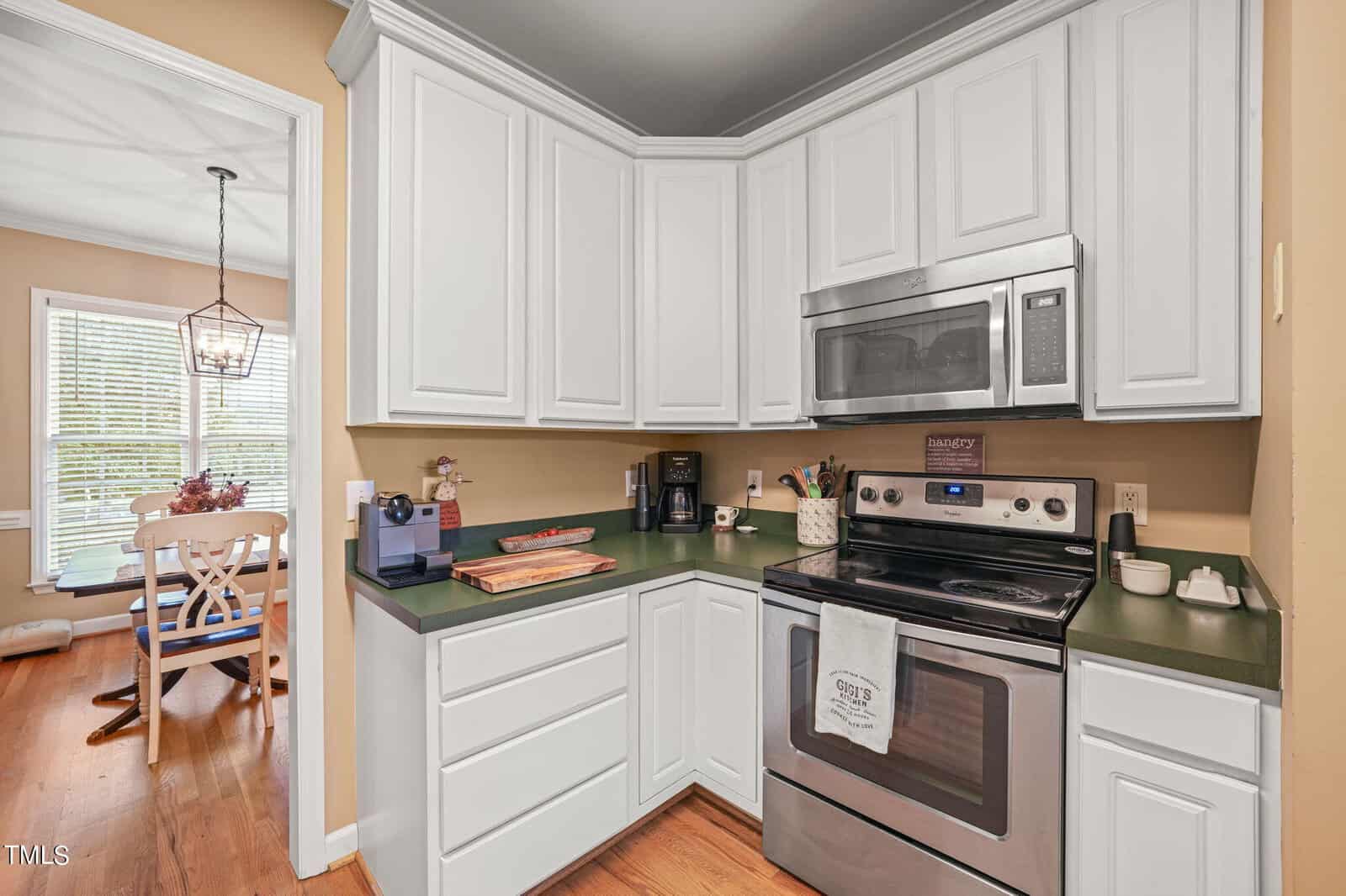 A kitchen with white cabinetry and green countertops features a stainless steel oven and microwave. A towel hangs on the oven door. A coffee maker and cutting boards are on the counter. A dining area with a window is visible in the background.