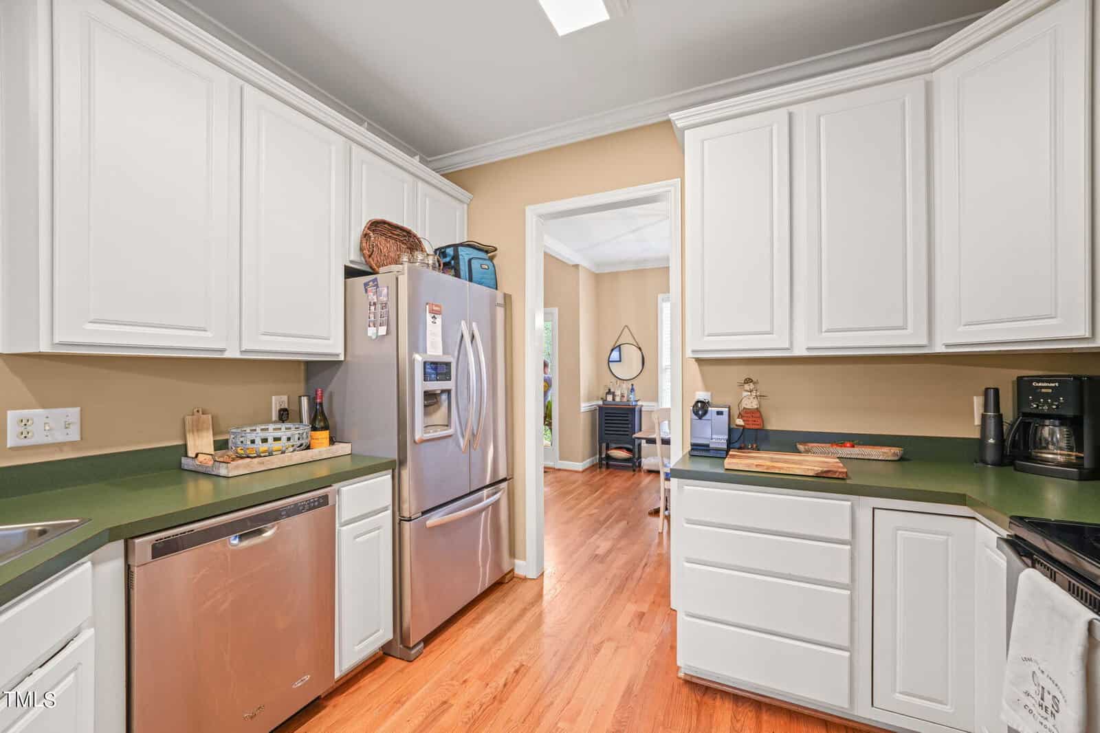 A modern kitchen with white cabinets and green countertops. It features stainless steel appliances, including a refrigerator and dishwasher. Wooden flooring complements the space. A door leads to an adjacent room with natural light.