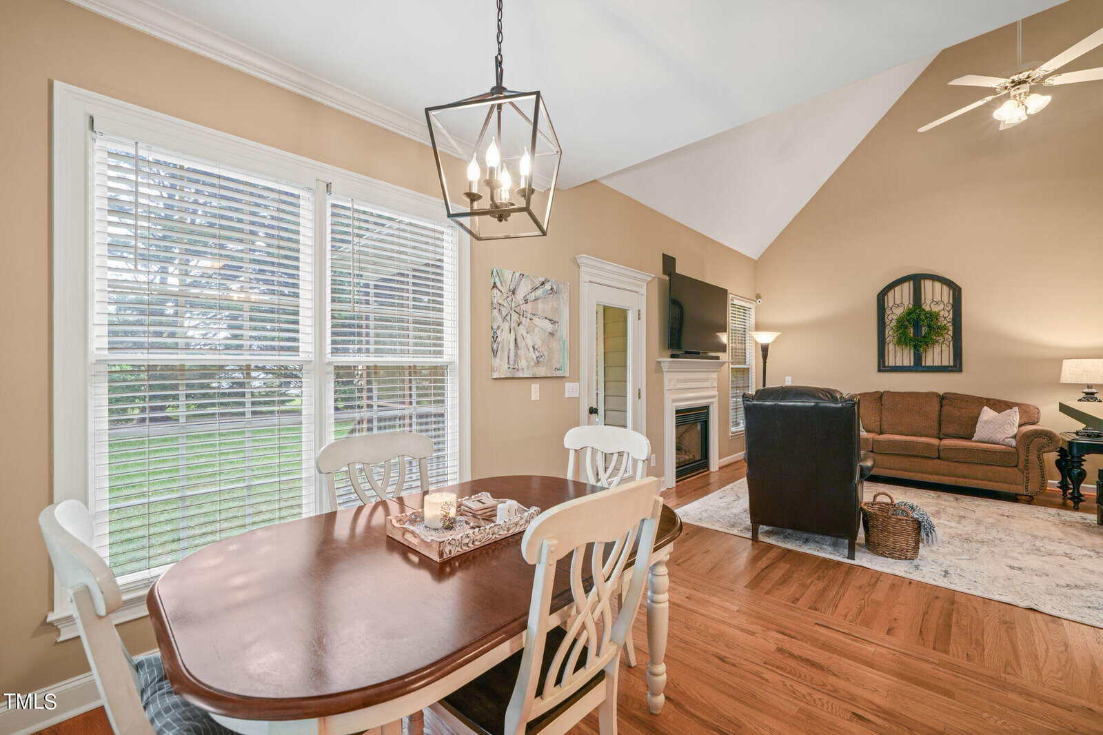 A cozy living and dining area with a wooden table and chairs, a gray sofa, and a fireplace. The room is well-lit by large windows, and a modern chandelier hangs above the table. A patterned rug and wall art add decorative elements.