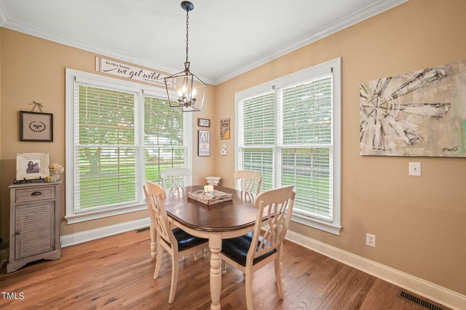 A cozy dining area with a wooden table and four chairs, surrounded by large windows with blinds. The walls are decorated with a clock and various wall art. A pendant light hangs above the table, and the flooring is wooden.