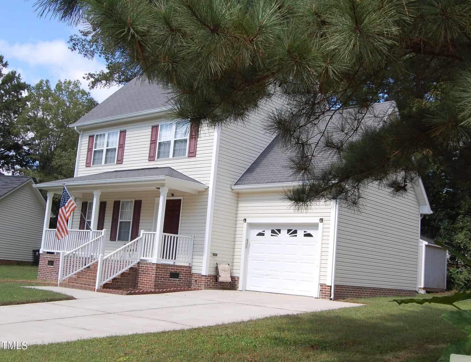 A two-story white house with a brick foundation sits proudly at 314 Williamsburg Drive in Creedmoor. An attached garage complements the home, while the front porch with white railings displays an American flag. It's surrounded by lush green grass and trees under a clear blue sky.