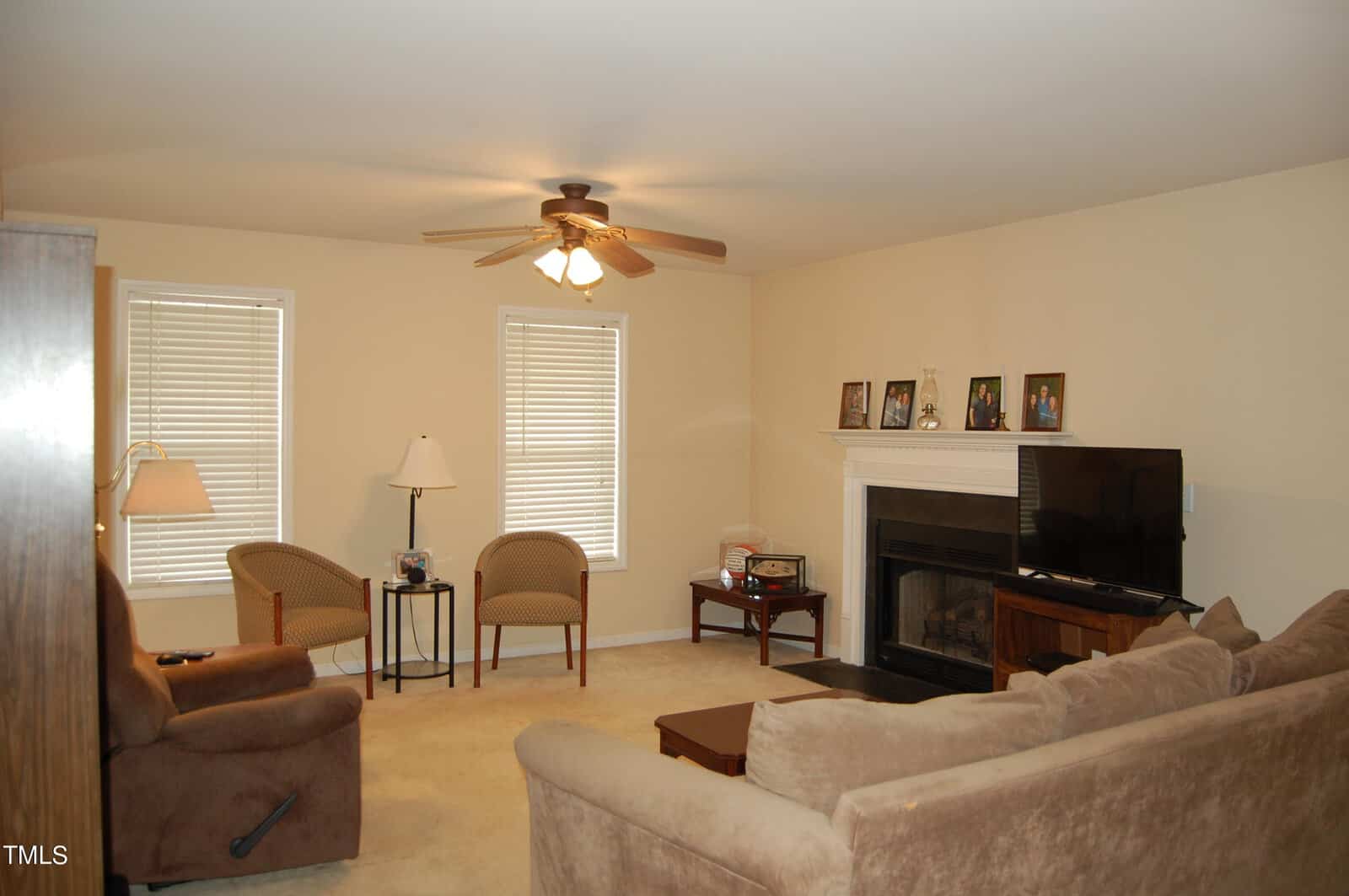 A living room with beige walls and carpet, featuring a ceiling fan, two windows with blinds, a sofa, an armchair, a recliner, and a wooden coffee table. A fireplace with family photos on the mantel and a flat-screen TV are present.