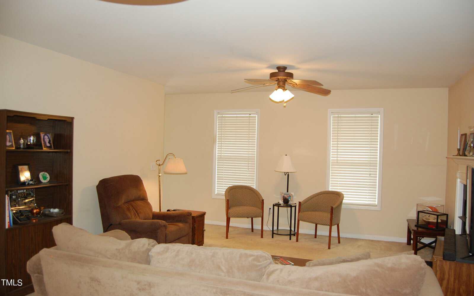 A cozy living room with a brown recliner, beige couch, two wicker chairs, and a small glass-top table. A bookshelf with photos and a fireplace with a TV are in the room. A ceiling fan and two windows add to the ambiance.
