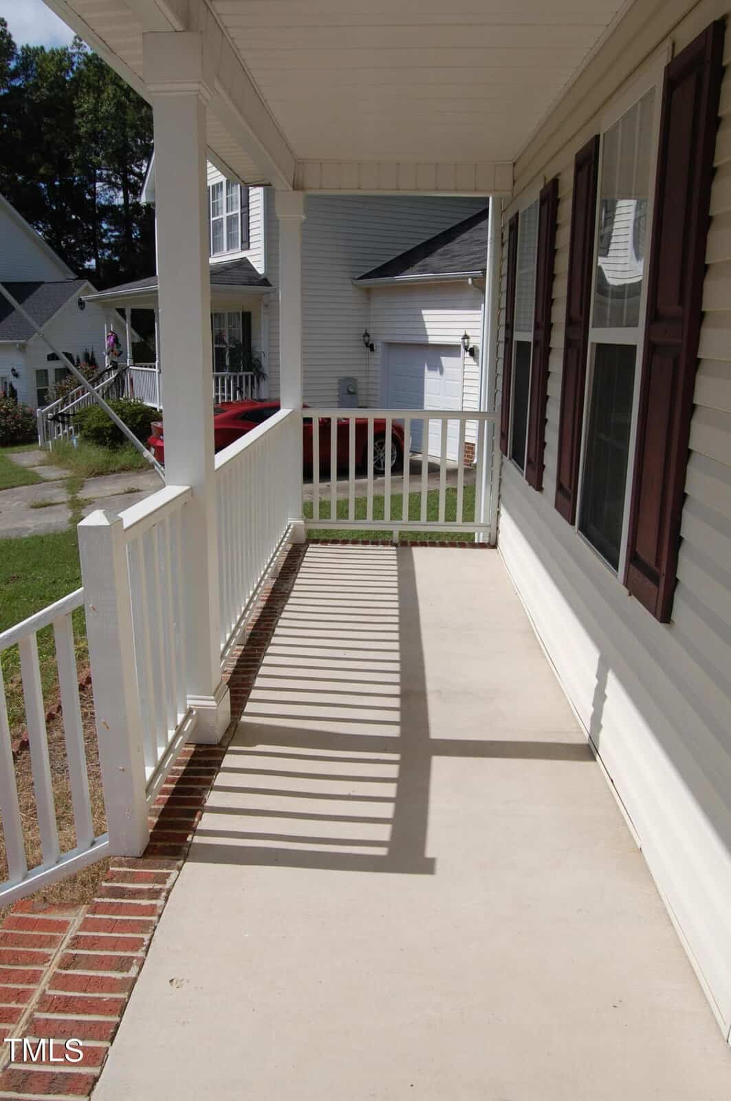 A covered porch with a white railing and brick trim. The porch leads to the front yard, with neighboring houses visible in the background. Sunlight casts shadows of the railing onto the concrete floor.