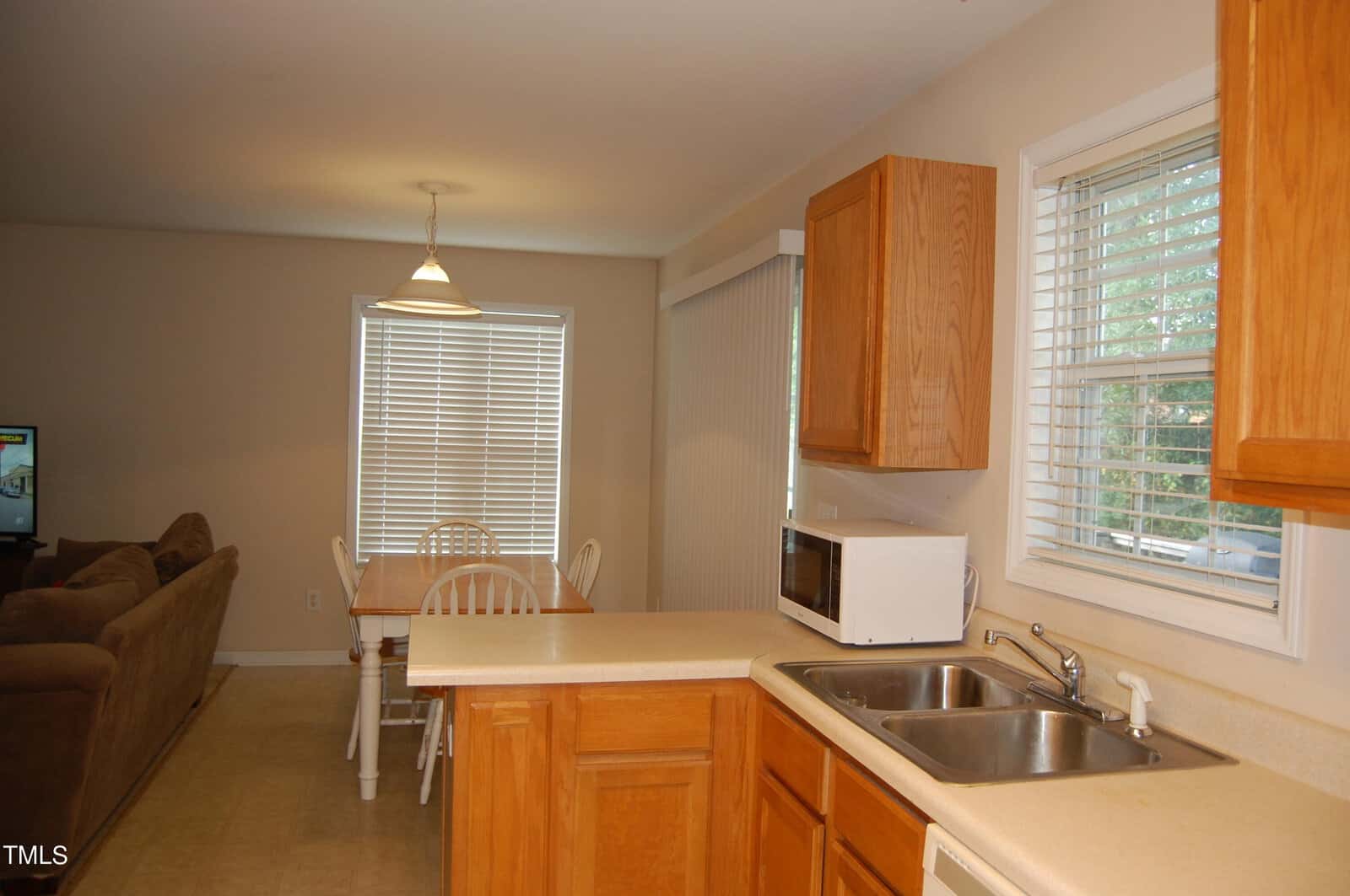 A kitchen with wooden cabinets, a double sink, and a white microwave on the counter. A dining area with a wooden table and chairs is near a window with blinds. A brown couch is visible in the living room area.