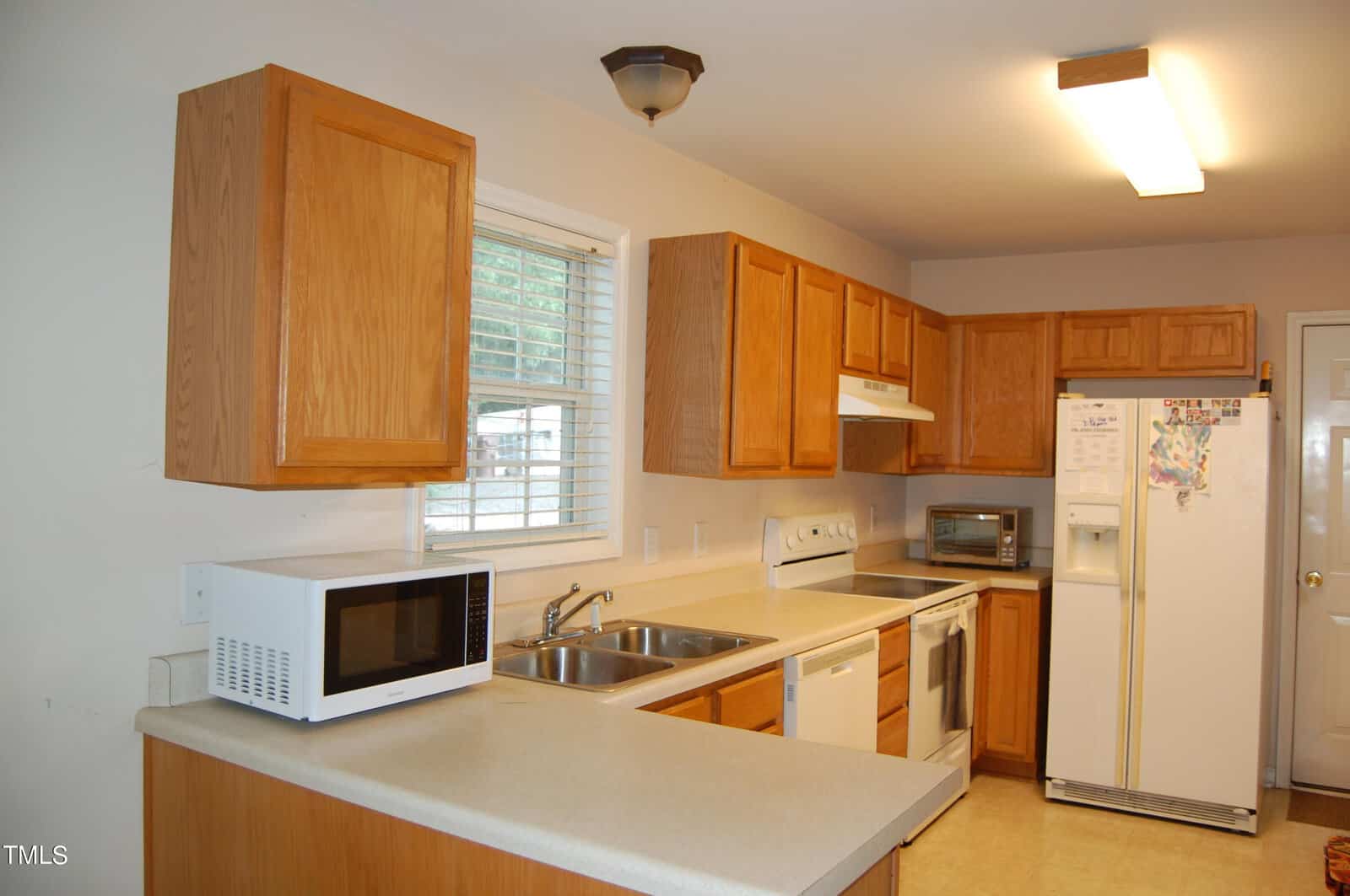 A kitchen with wooden cabinets, a white refrigerator, and a microwave on the countertop. There is a stainless steel sink under a window, a stove, and a toaster oven. The room is lit with ceiling fixtures.