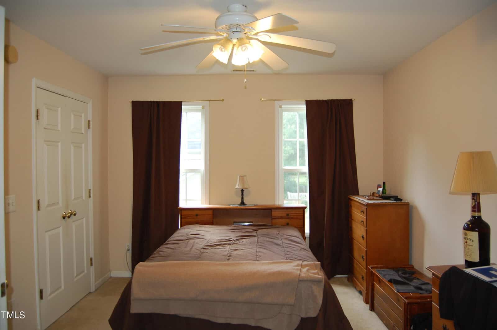 A bedroom with a ceiling fan, brown curtains, and beige walls. It features a bed with a brown and beige comforter, two windows, a nightstand with a lamp, and wooden dresser furniture. Natural light enters through the windows.